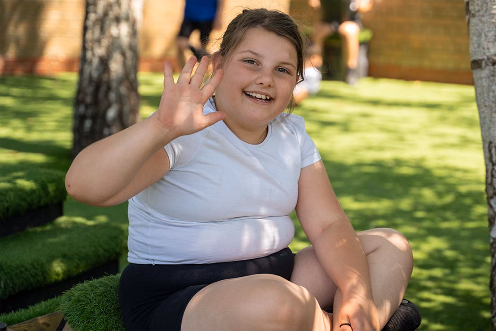 Pupil sitting on grass steps, smiling and waving at the camera.