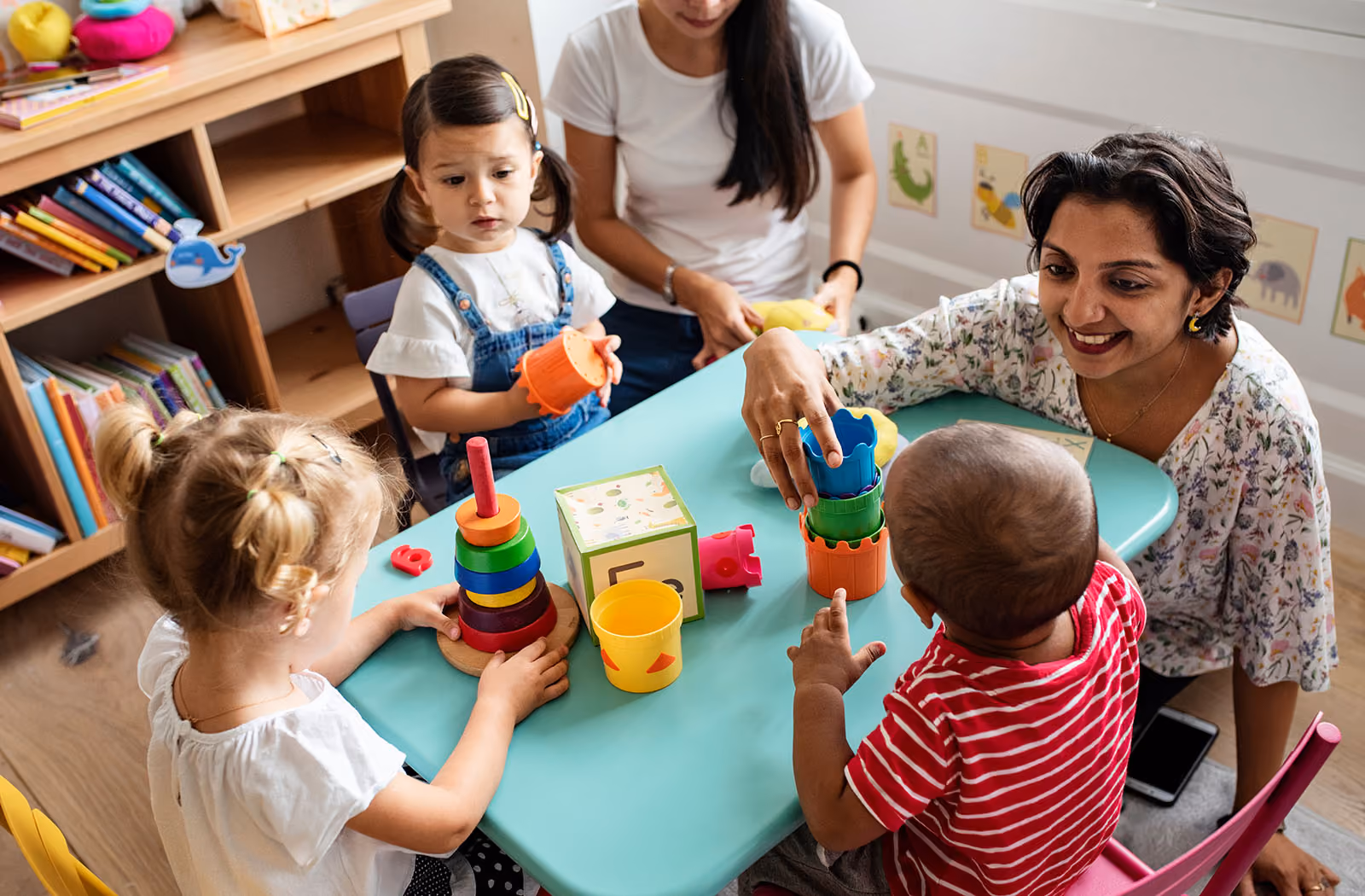 Nursery staff and young children playing with stacking toys at a table.