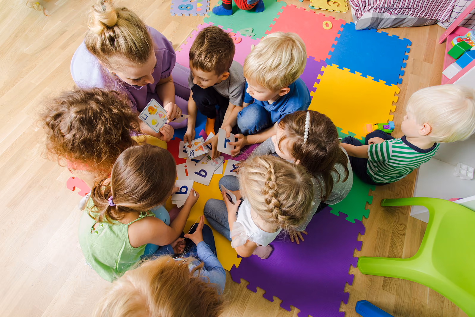 Overhead view of nursery staff and young children playing with letter cards on a colourful floor mat.