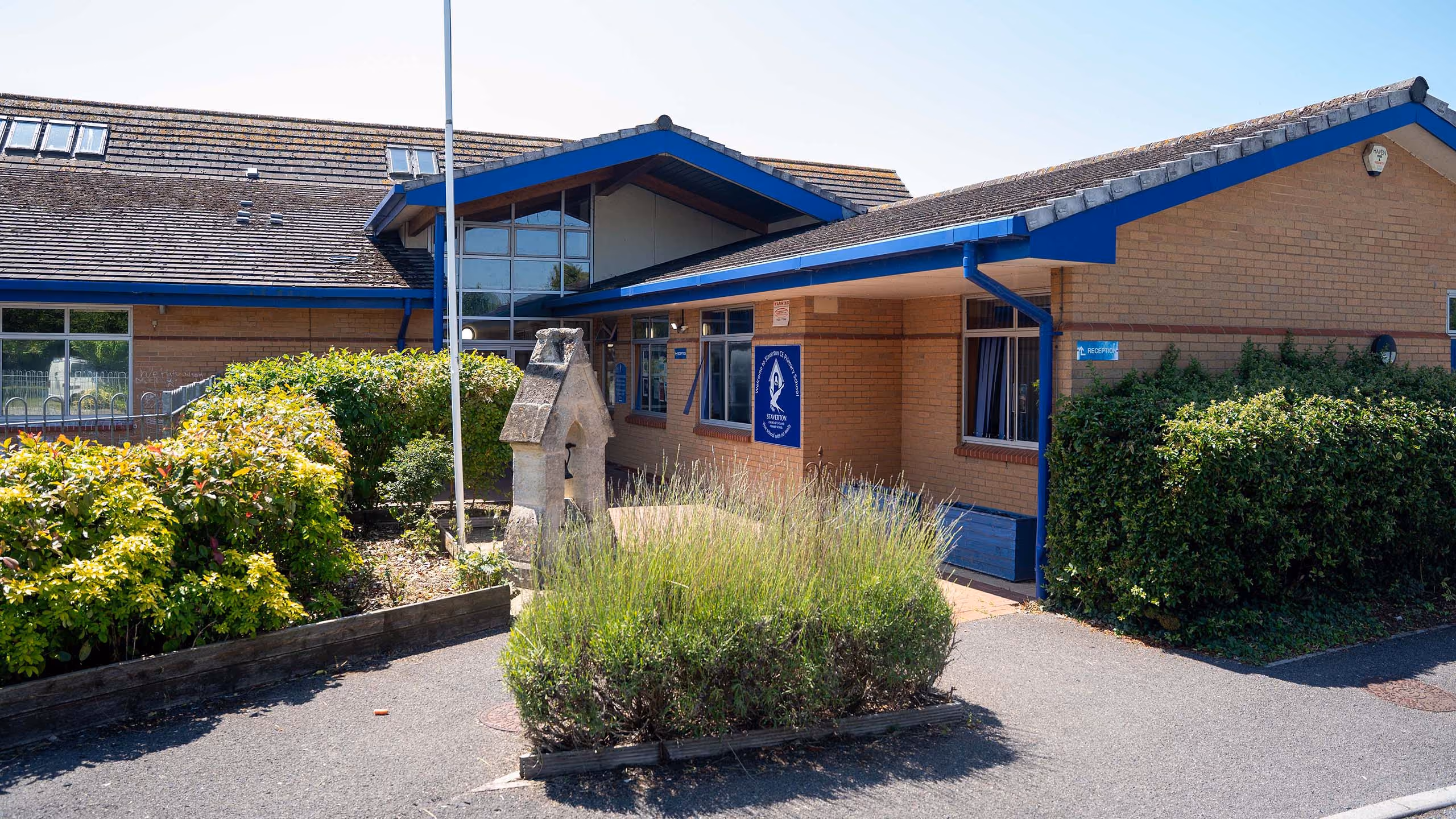 Brick school building with blue trim, bushes, and a stone bell structure in front on a sunny day.