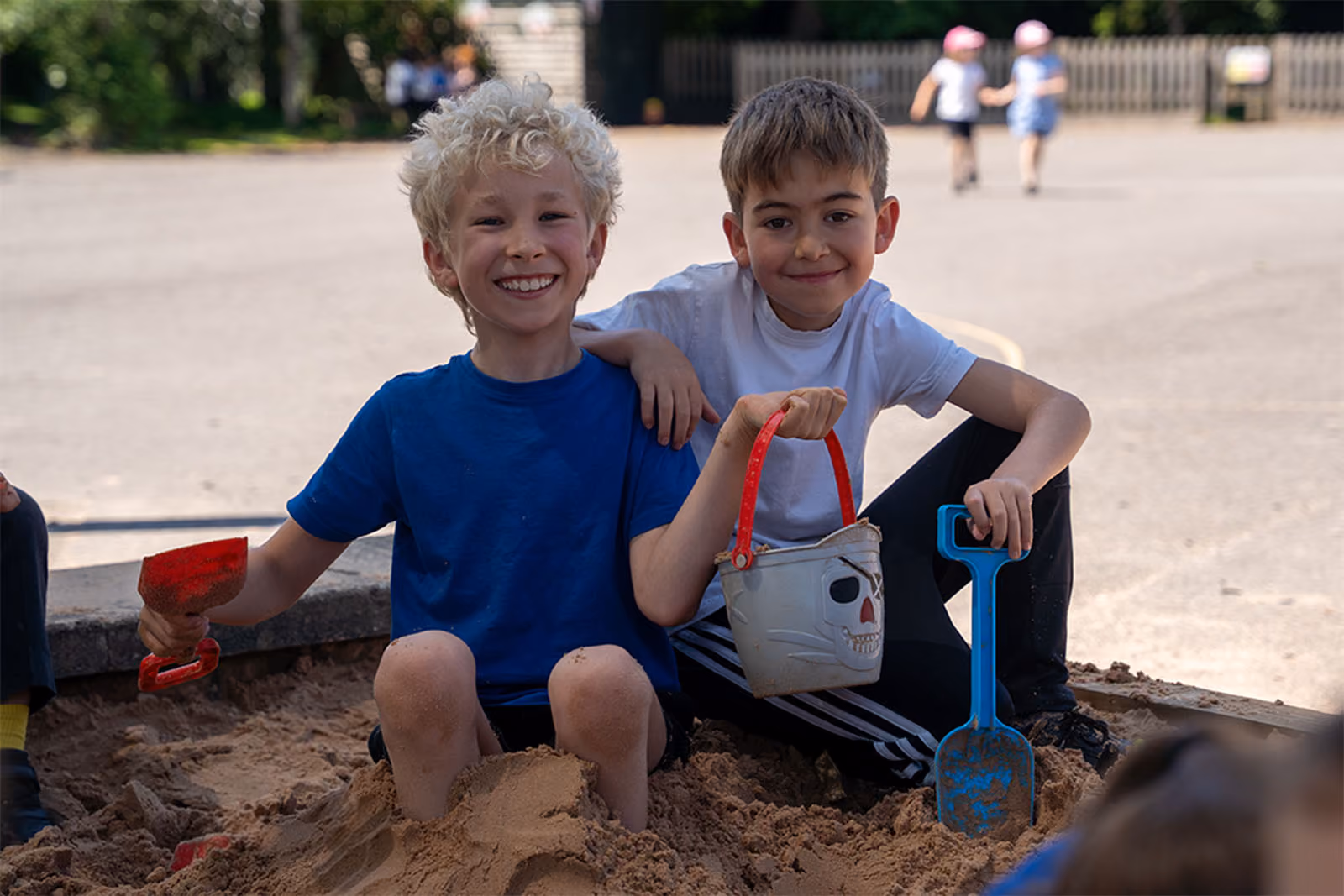 Two boys sitting and smiling in a sandbox holding a skull bucket and blue shovel, outdoors on a sunny day.