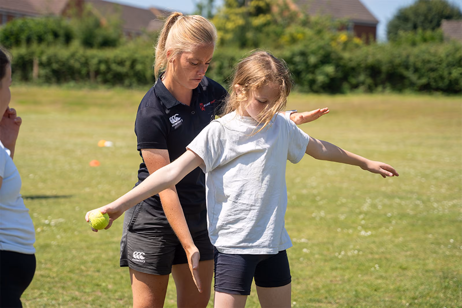 Adult woman coaching young girl with outstretched arms holding tennis ball outdoors on grass field.