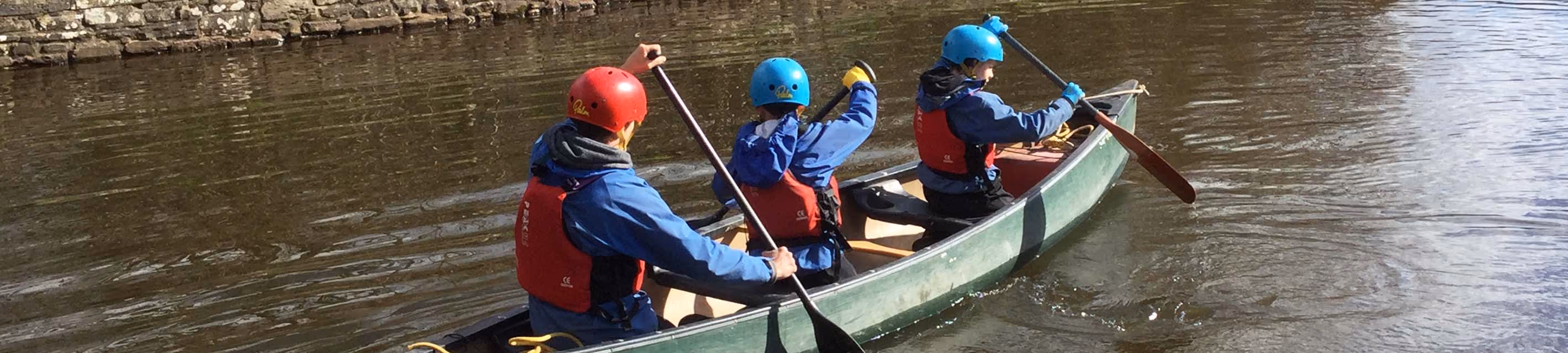 Three pupils in helmets and life jackets paddling a canoe on a river during a residential trip.
