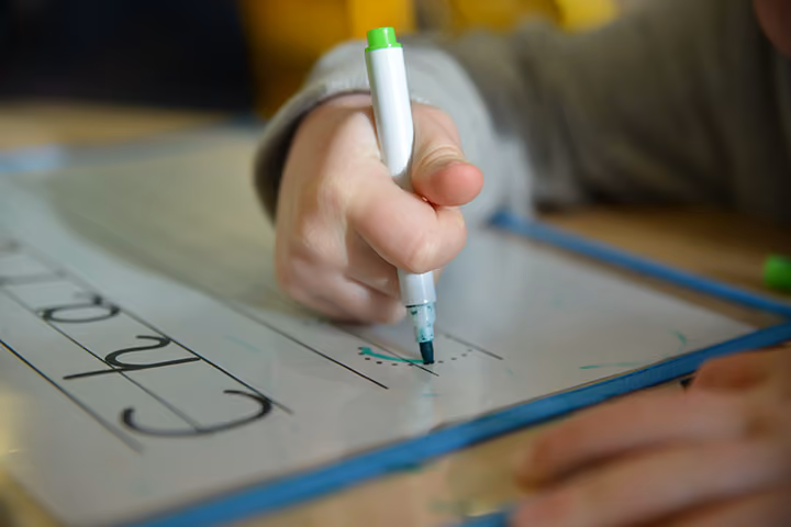 Close-up of a child's hand practising handwriting with a marker on a whiteboard.