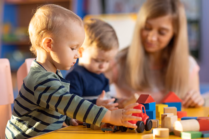 Toddler playing with toy vehicles and blocks at a table with an adult and another child.