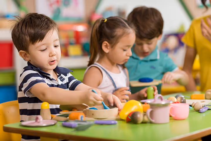 Young children playing with wooden toy food and kitchen items at a table.