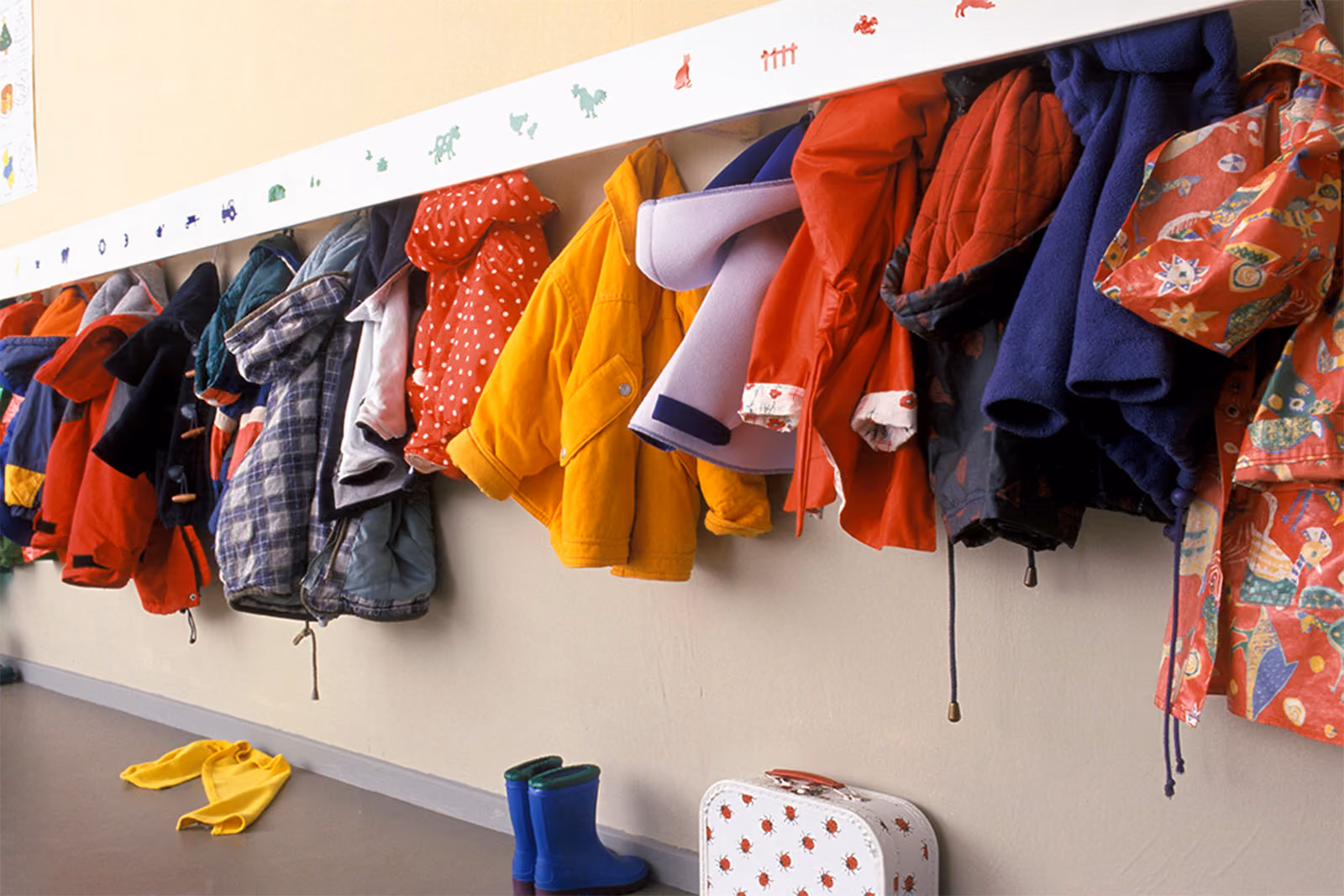 Row of children's coats hanging on pegs above wellington boots and a lunchbox.