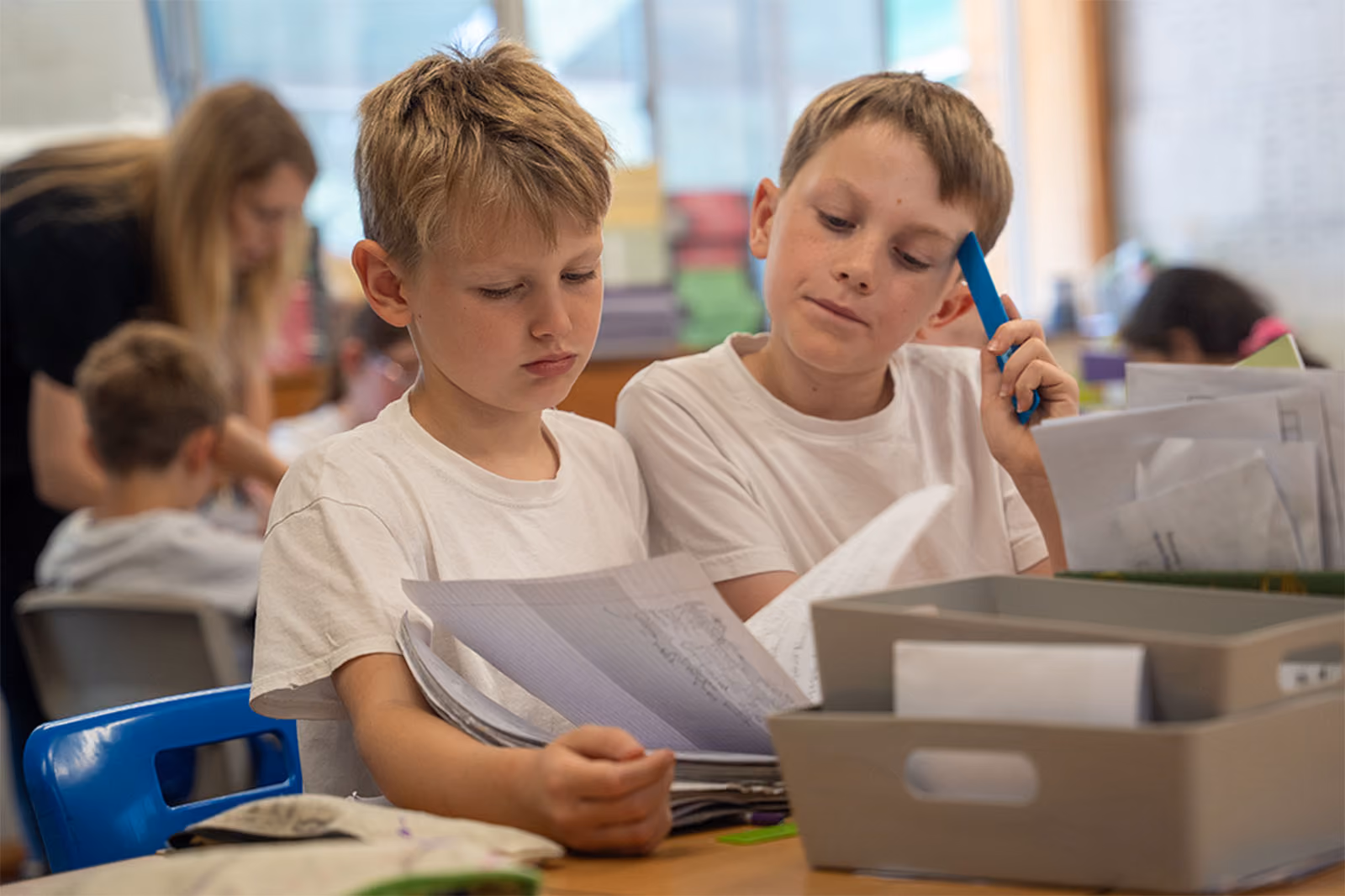 Two boys in white shirts reviewing papers together at a classroom desk with supplies.
