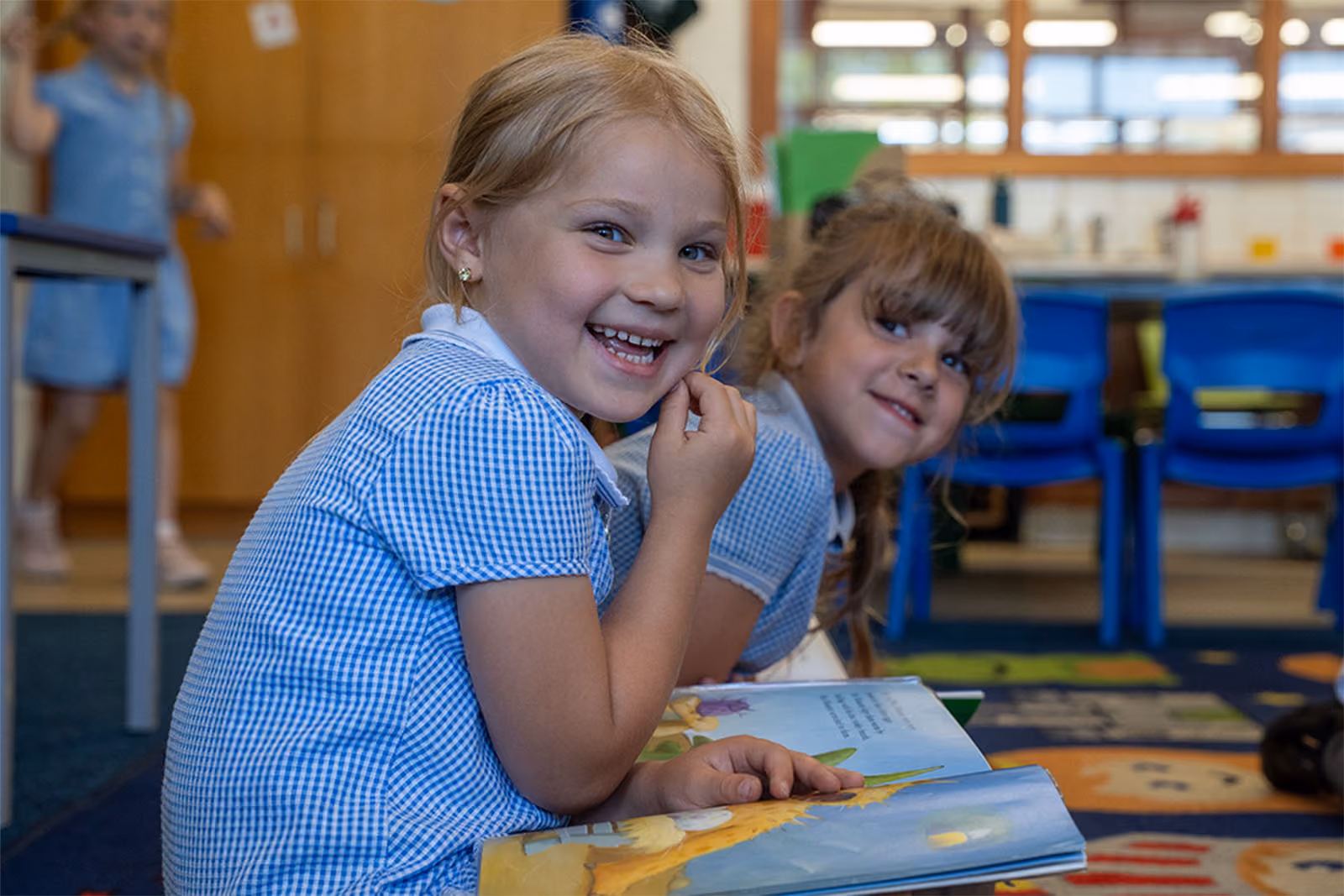 Two young girls in blue checkered dresses smiling while sitting indoors, one holding a book.