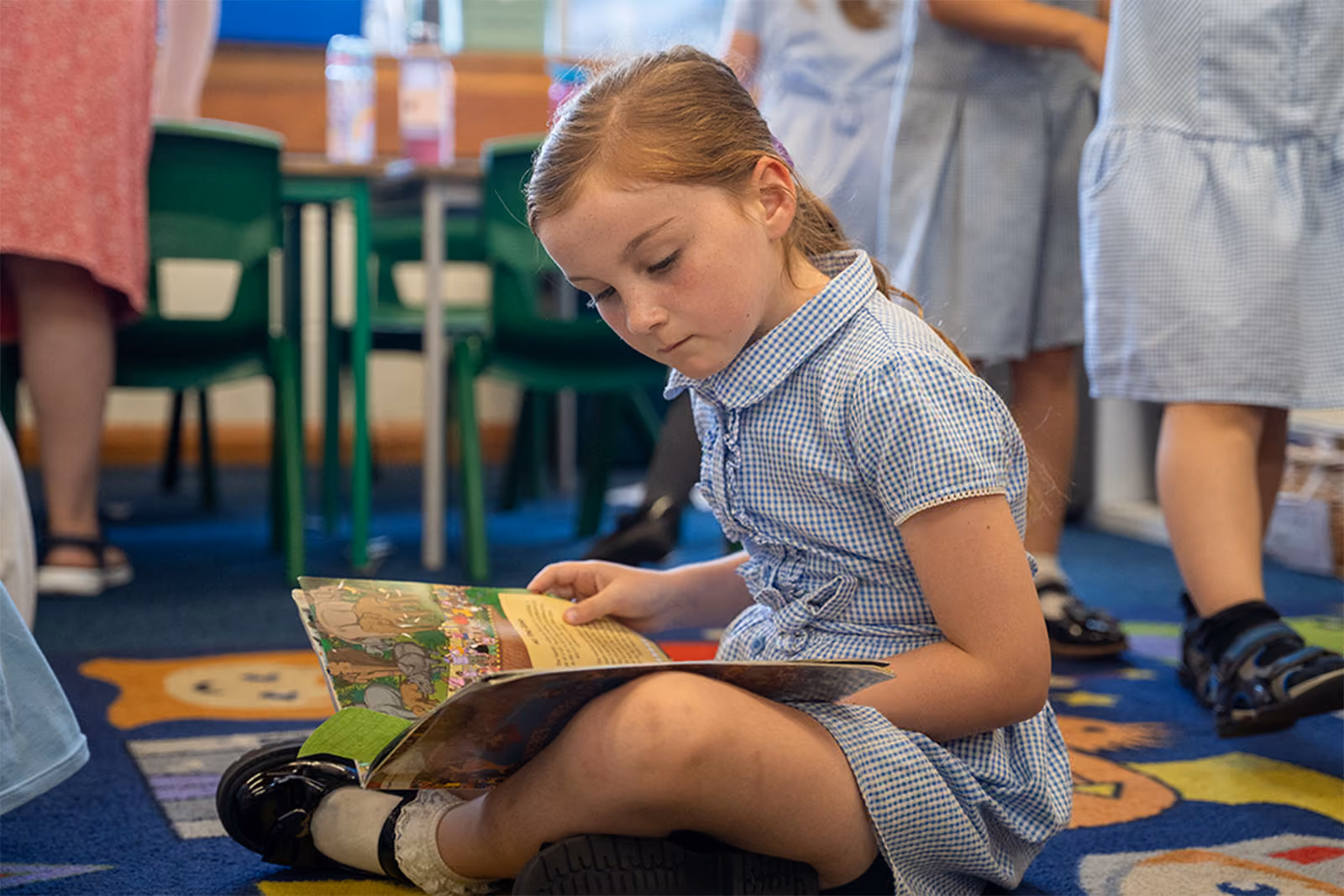 Young girl in a blue checkered dress sitting on carpet reading a book in a classroom.