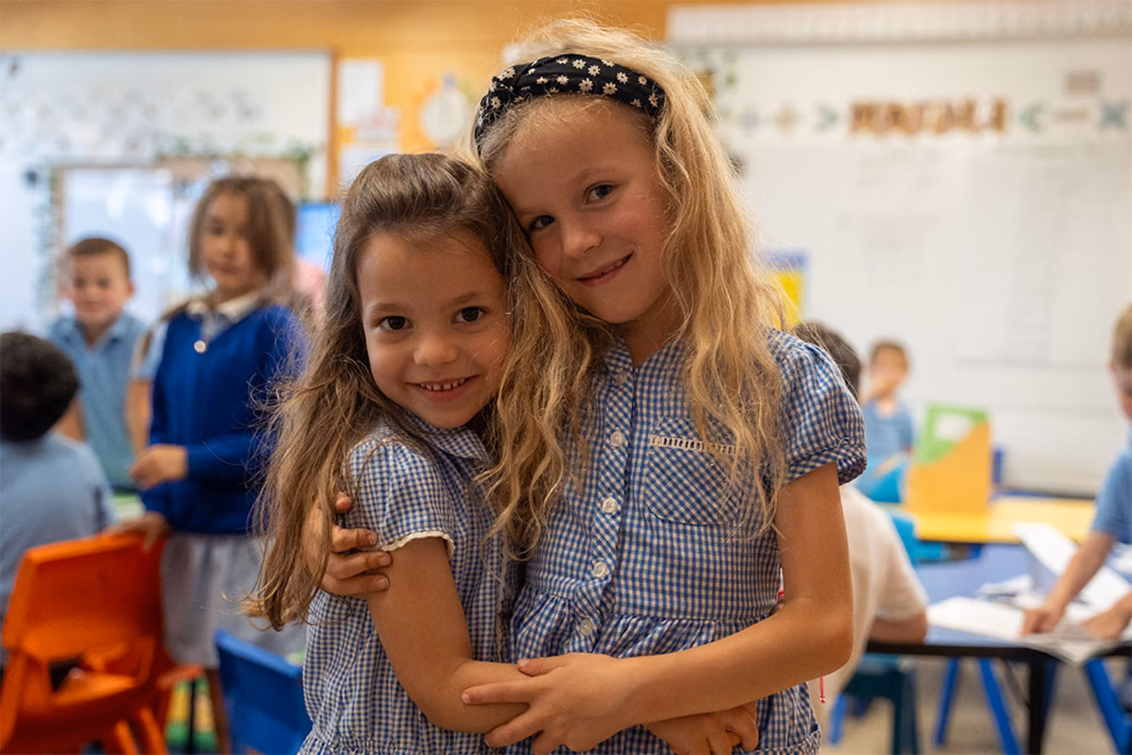 Two smiling pupils in school uniform hugging in a classroom.