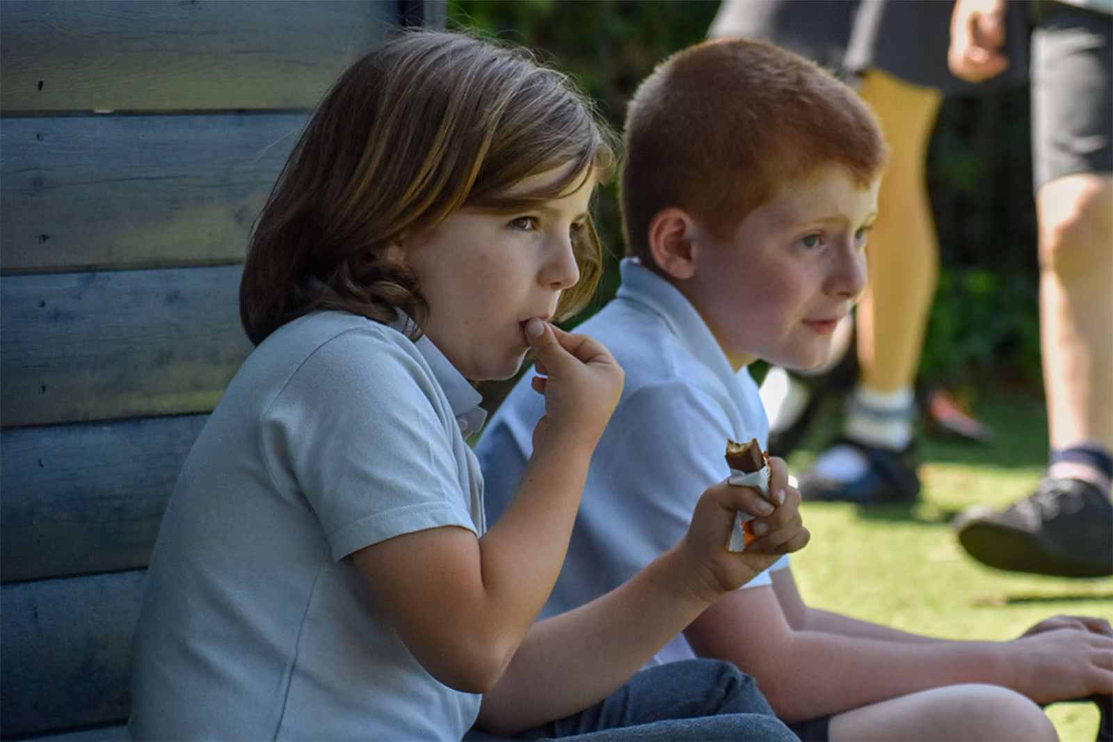 Two pupils sitting outside, one eating a snack, watching something off camera.