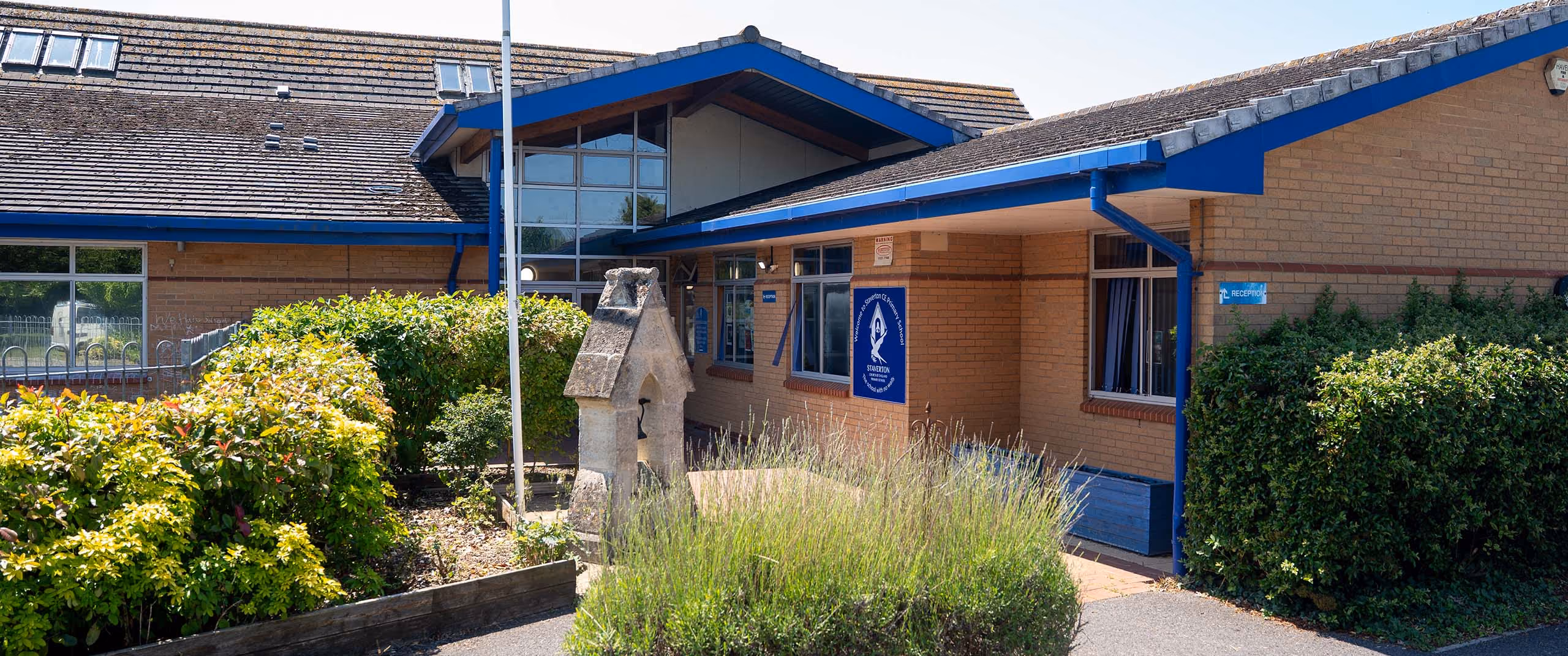 Staverton Primary School building with blue trim, bushes, and a stone bell structure in front on a sunny day.