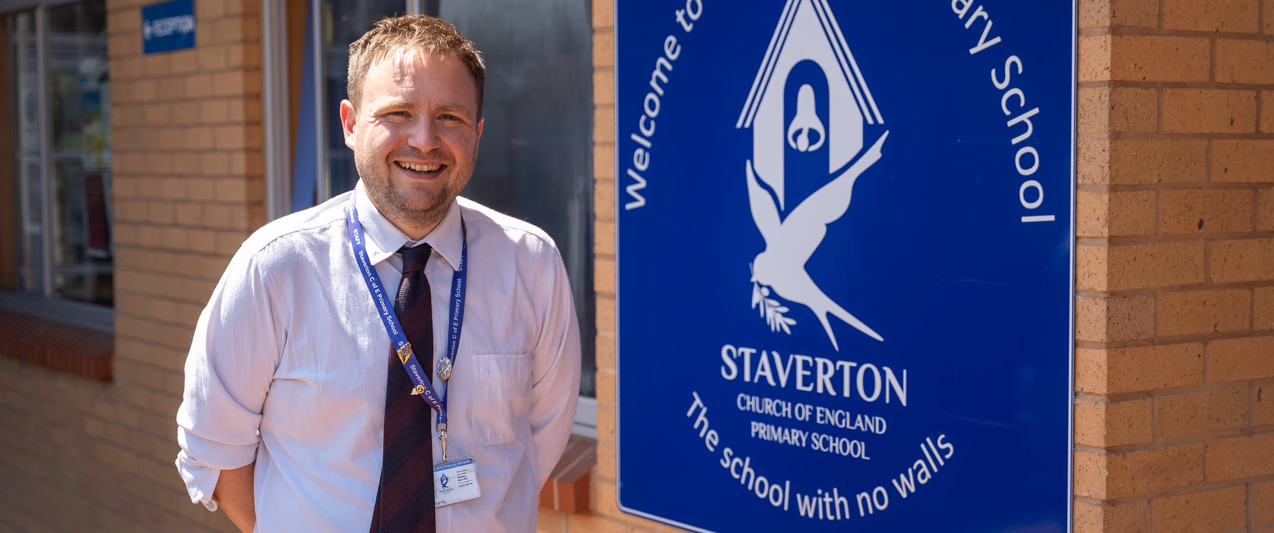 Head teacher in a white shirt and tie standing beside the Staverton CE Primary School sign.
