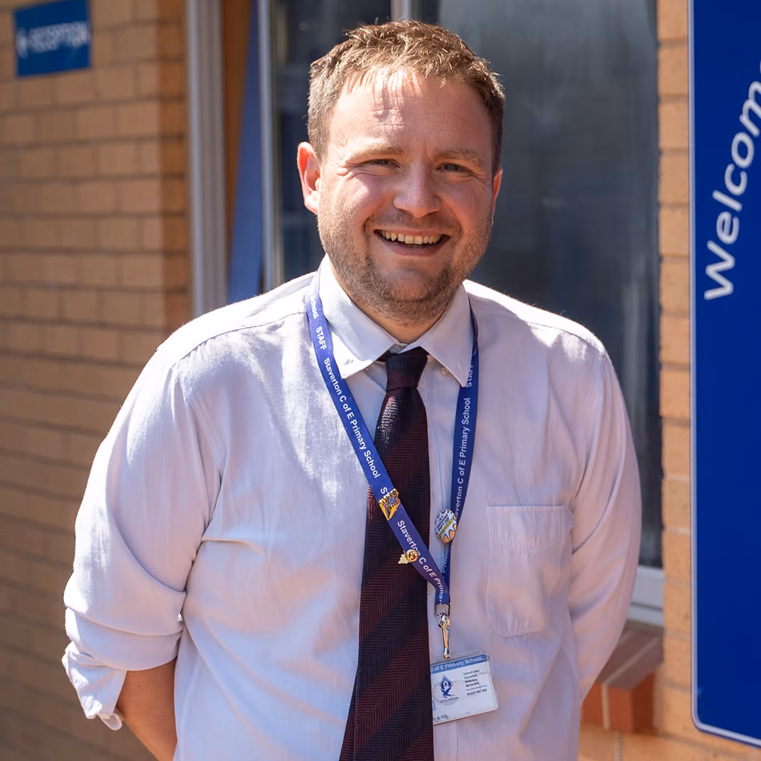 Head teacher in a white shirt and tie standing beside the Staverton CE Primary School sign.