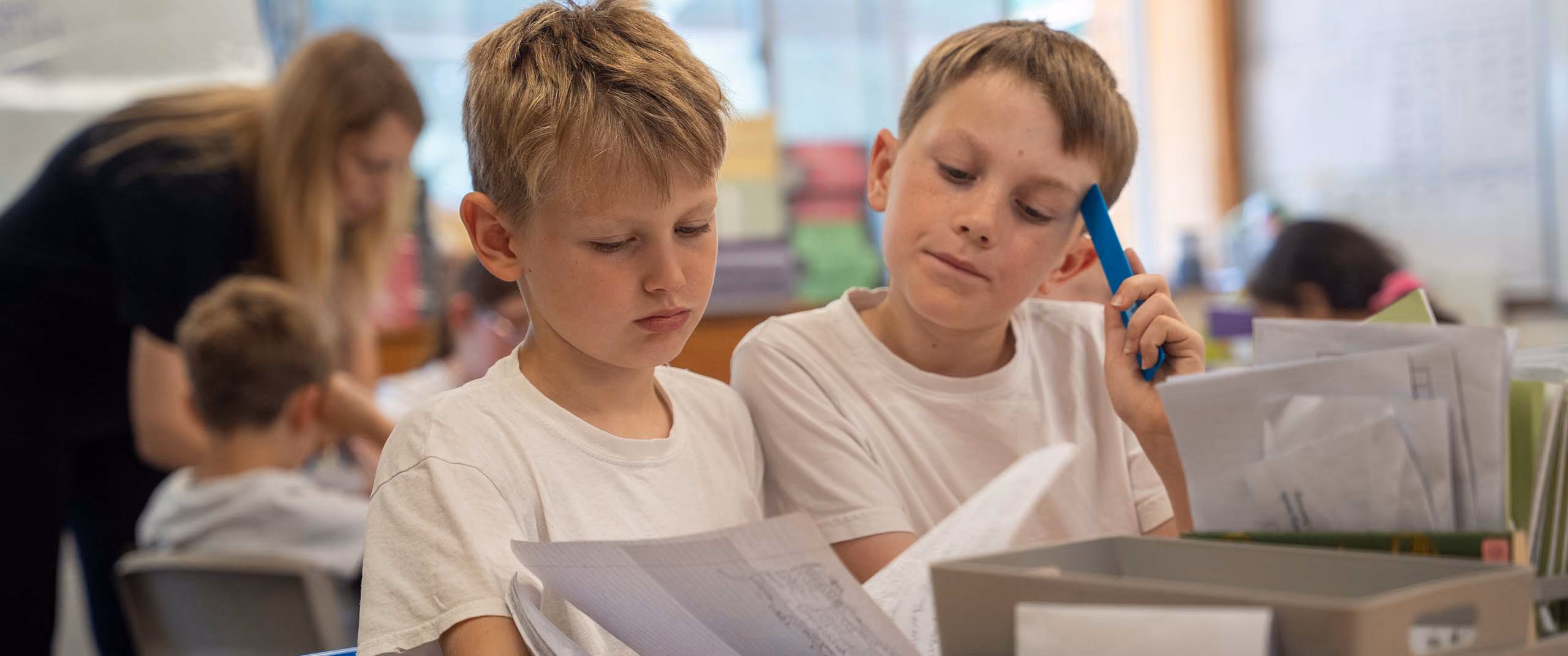 Two boys in white shirts reviewing papers together at a classroom desk with supplies.
