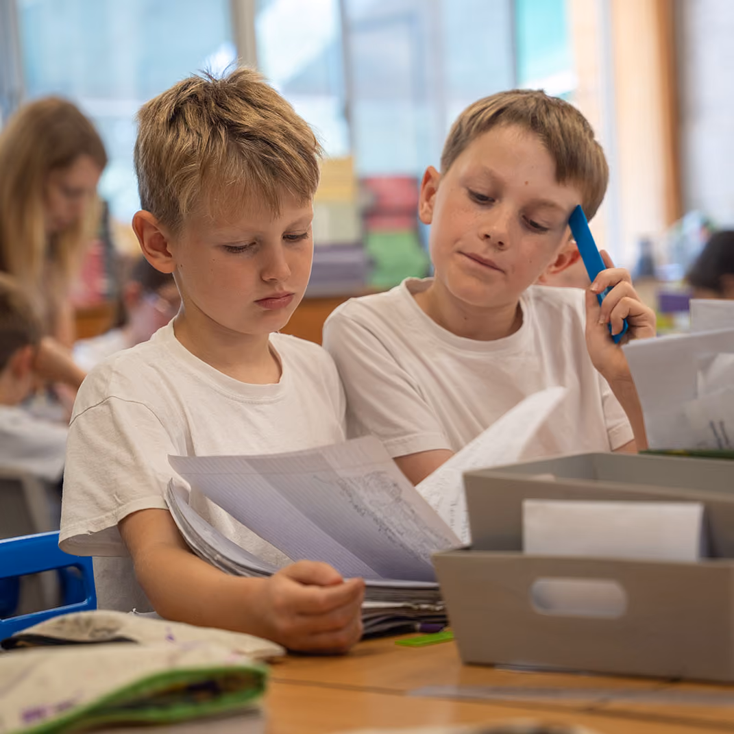 Two boys in white shirts reviewing papers together at a classroom desk with supplies.