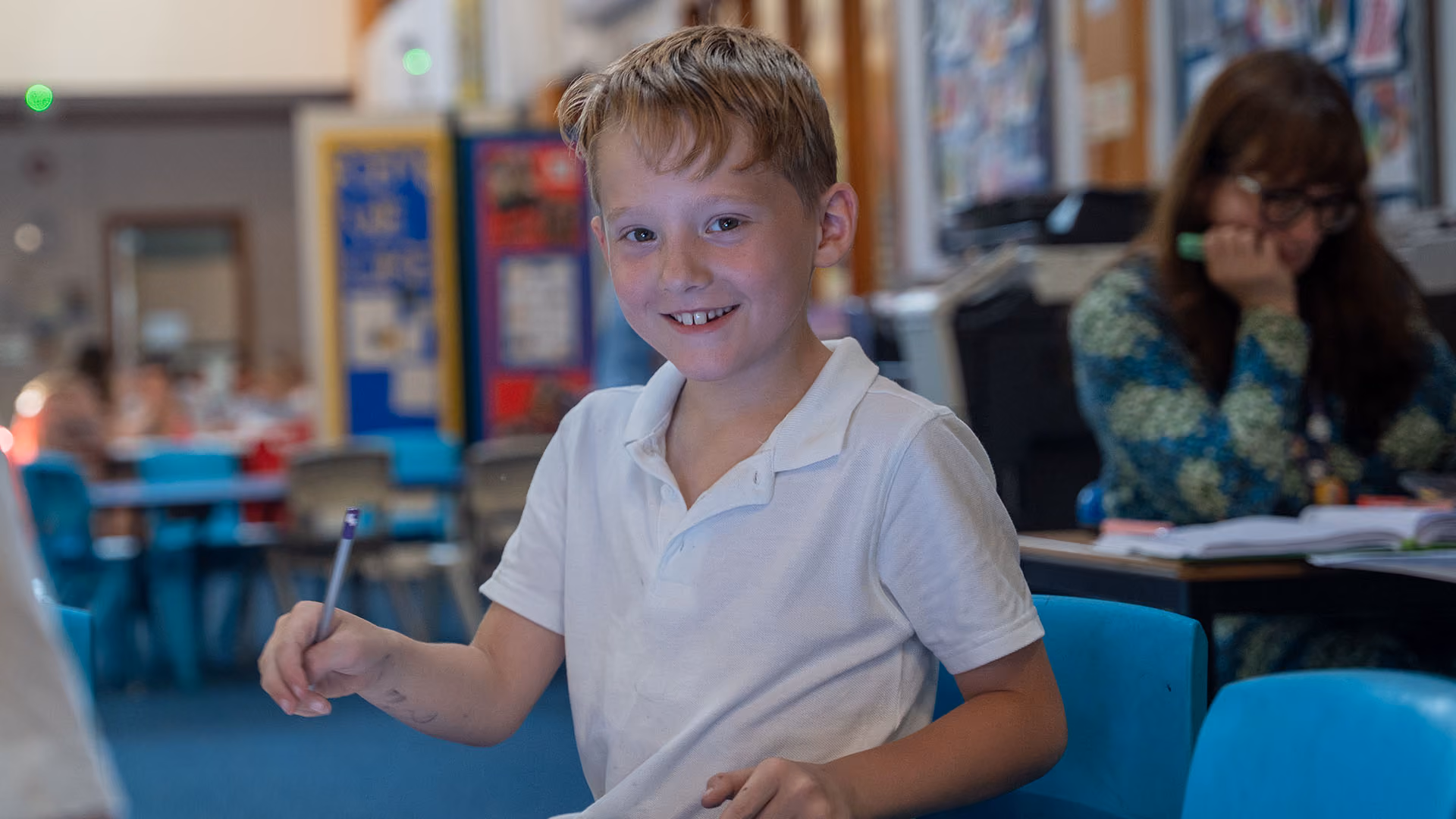 Smiling pupil holding a pencil in a classroom, with a teacher working at a desk behind.