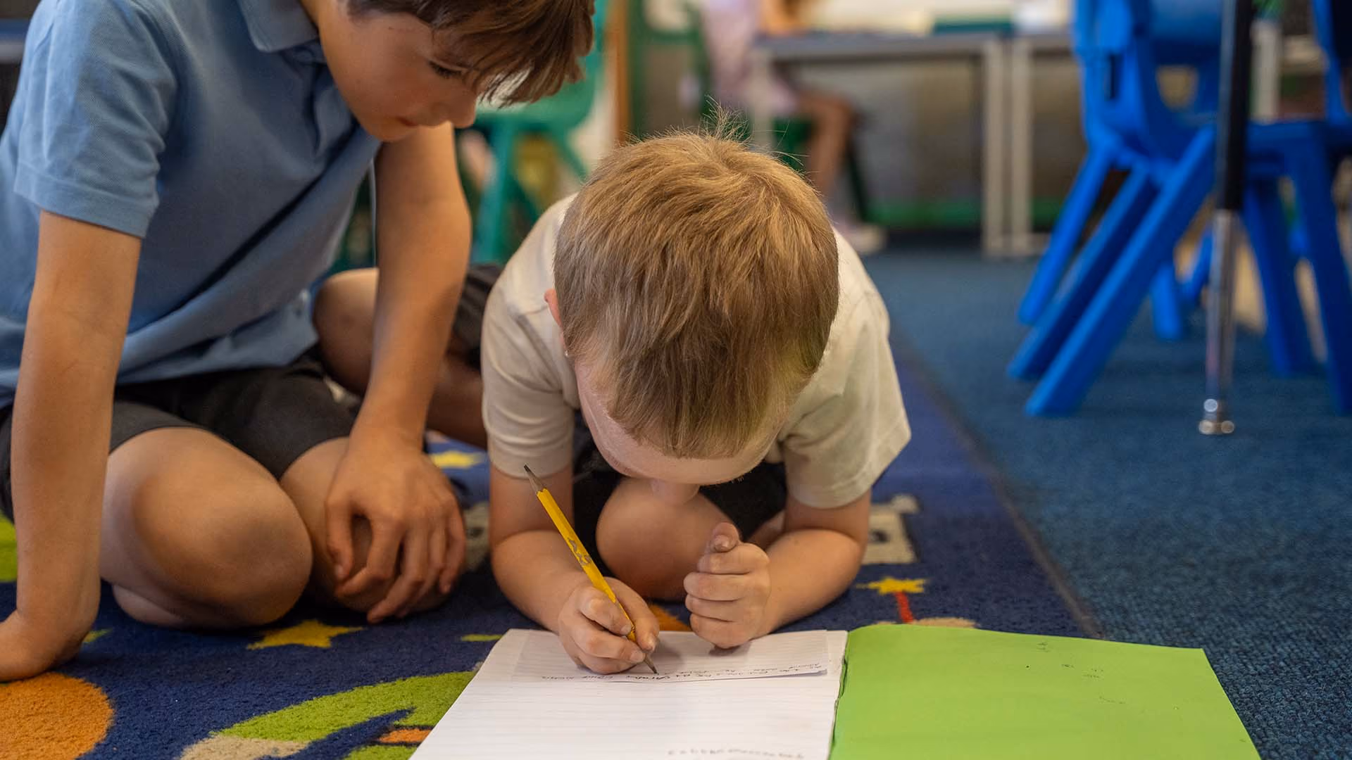 Two pupils on the classroom floor, one writing in an exercise book while the other watches.