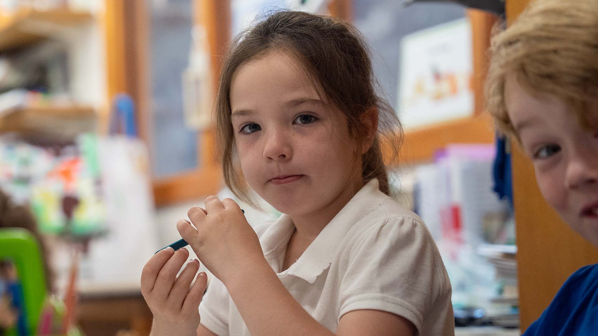 Pupil holding a pen looking at the camera, sitting beside another pupil in a classroom.