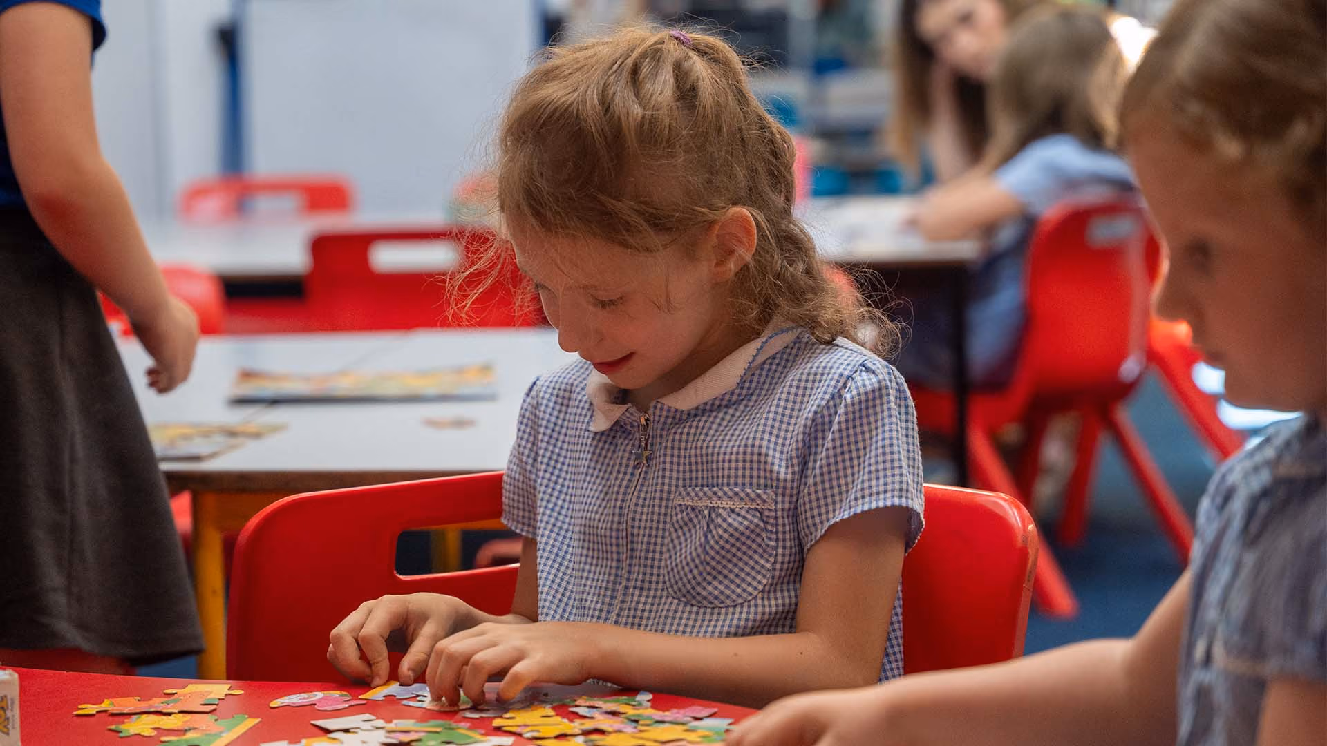 Young girl in a checkered dress assembling a colourful puzzle at a red table in a classroom.