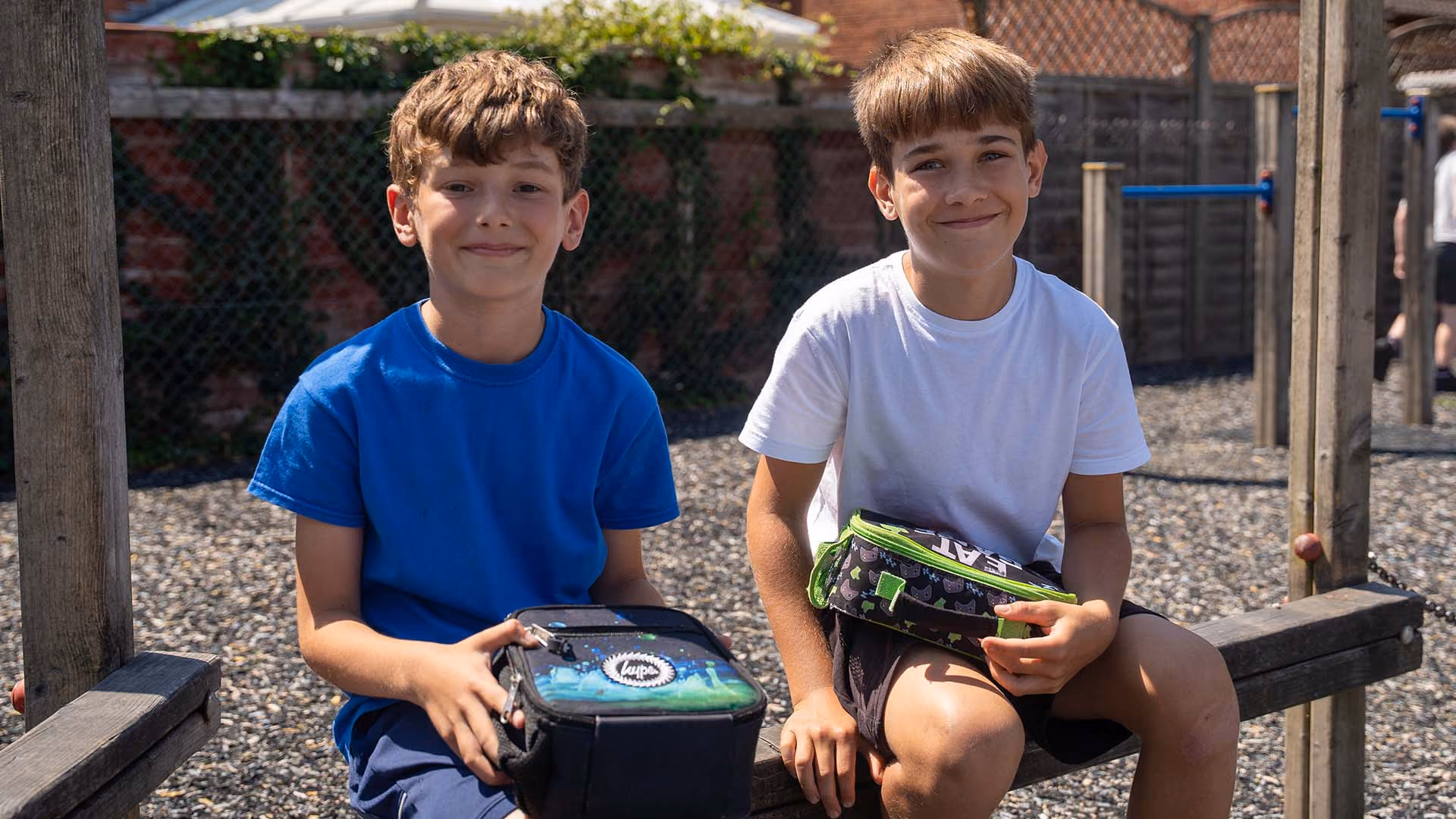 Two smiling pupils sitting on a wooden bench outside, each holding a lunch bag.
