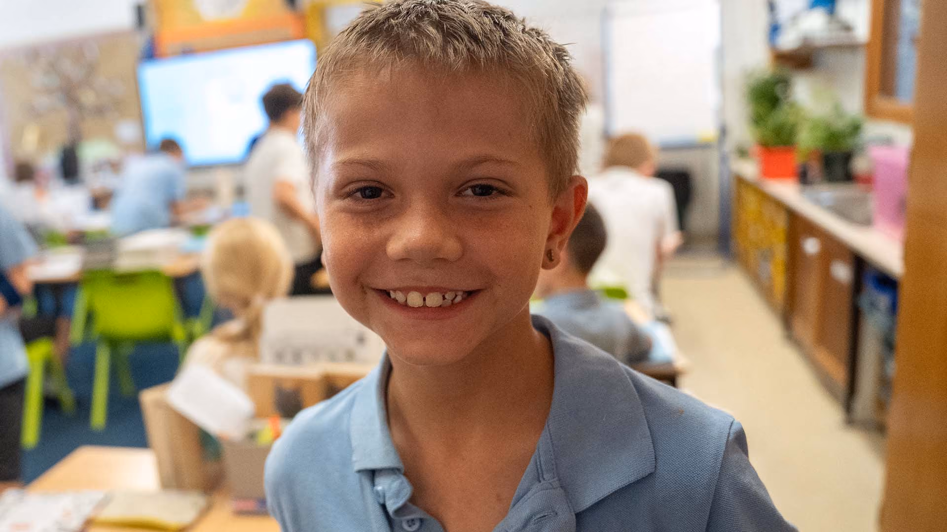 Smiling pupil in a light blue polo shirt in a classroom with other pupils working behind.