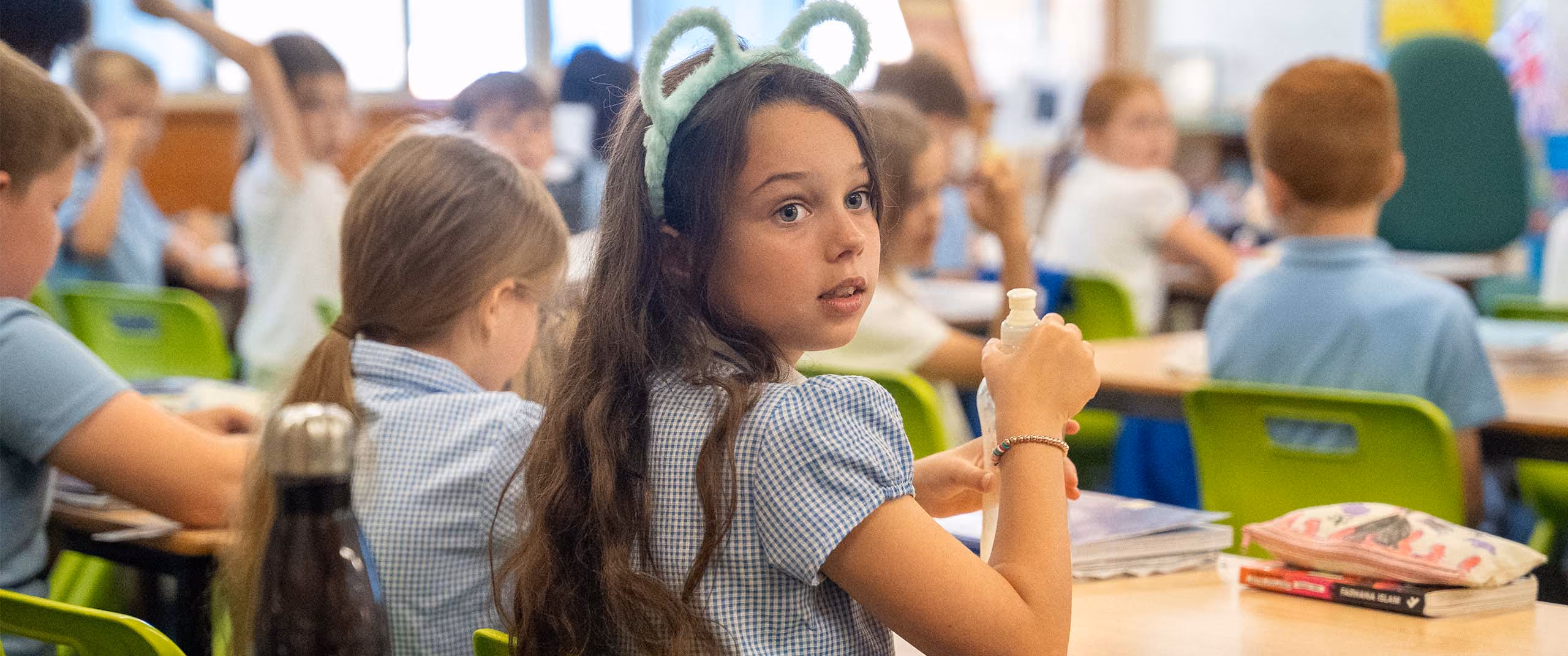 Pupil in a gingham dress turning to look at the camera in a busy classroom.