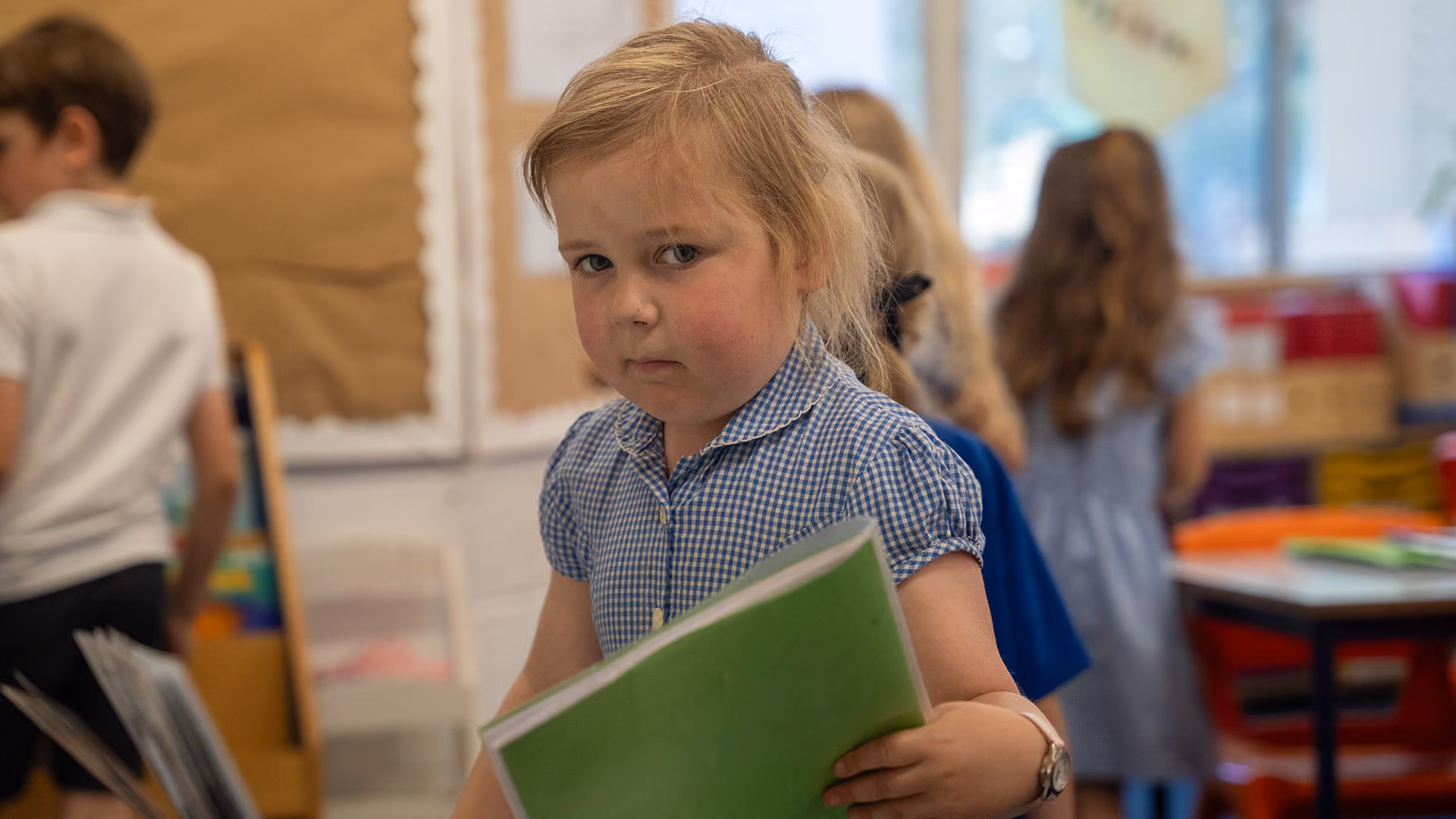 Pupil in a gingham dress holding a green exercise book in a busy classroom.