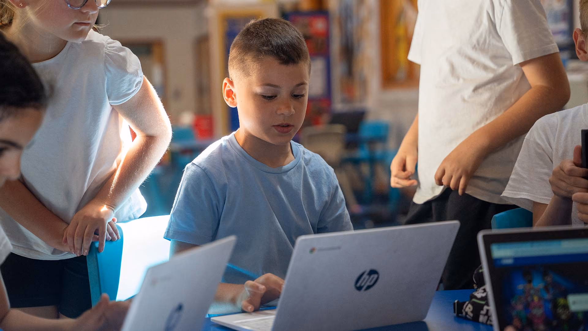 Young boy using an HP laptop at a table surrounded by other children in a classroom.