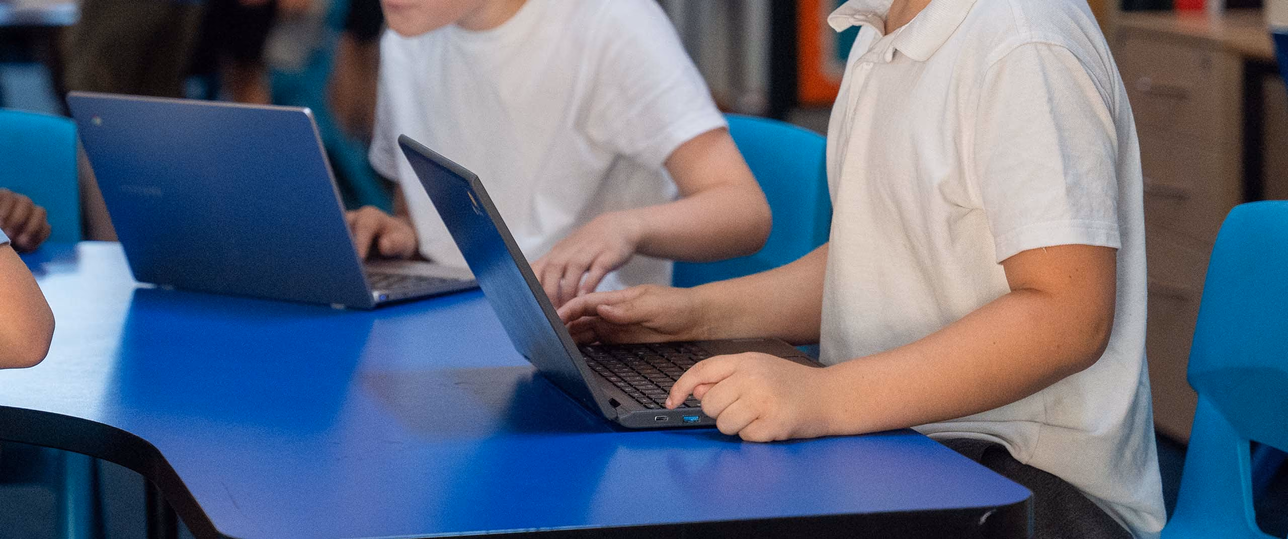 Two pupils typing on laptops at a blue classroom table.