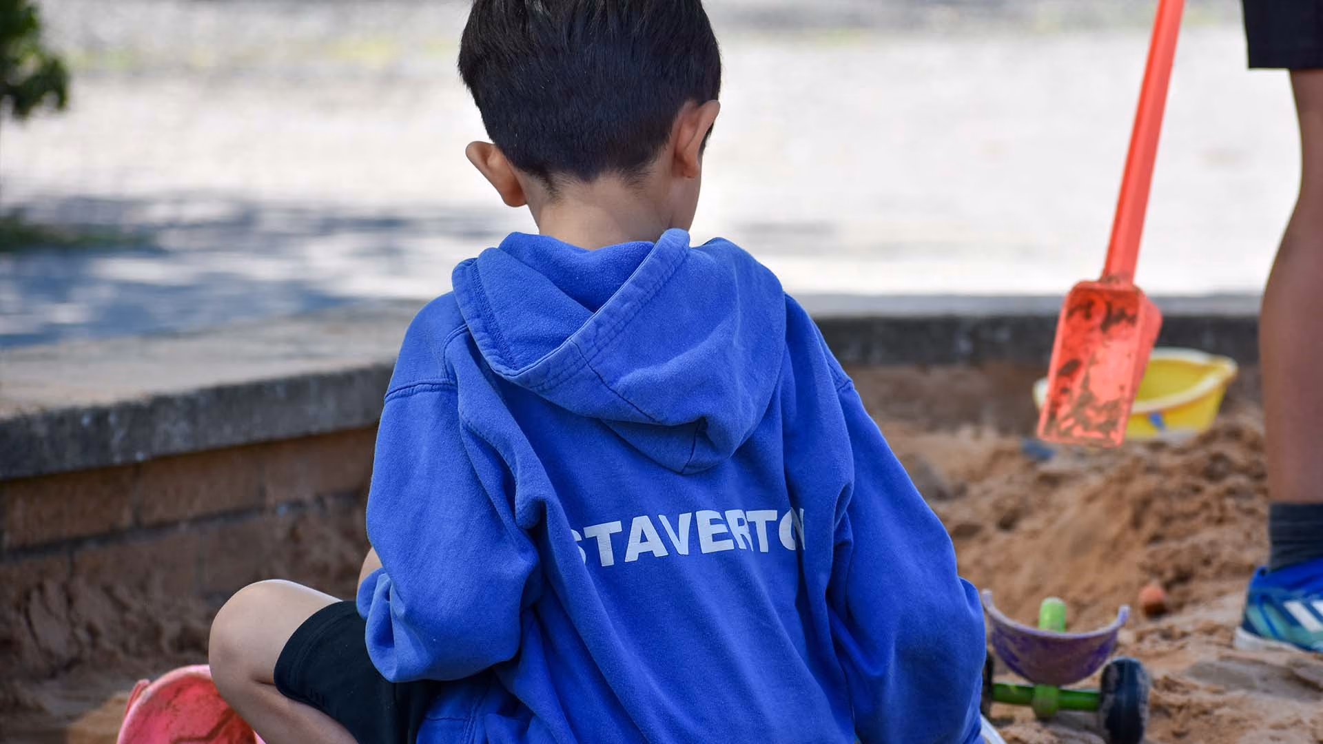 Child in a blue Staverton hoodie playing in a sandbox with toys, viewed from behind.