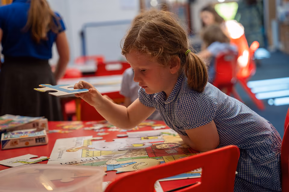 Pupil in a gingham dress fitting a piece into a jigsaw puzzle at a table.