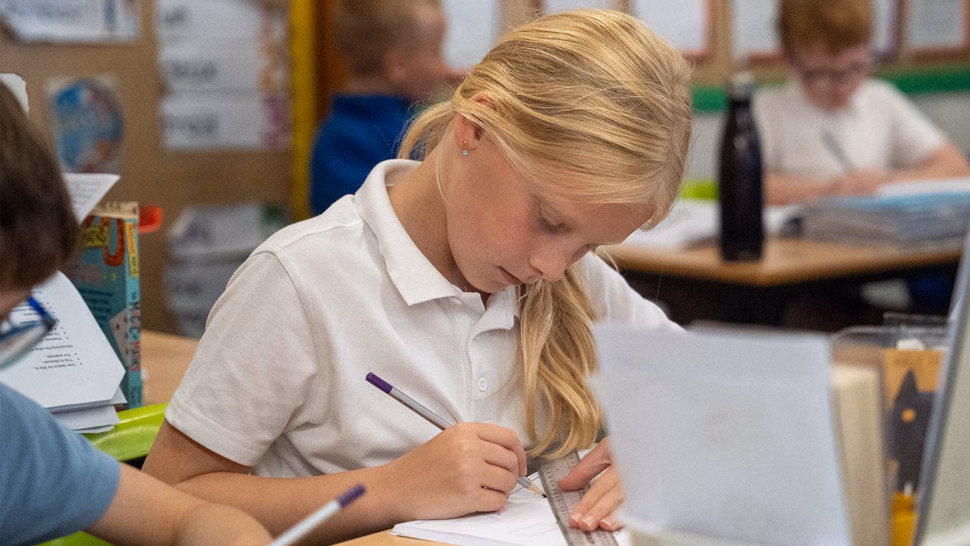 Pupil in school uniform using a ruler and pencil to write in an exercise book at her desk.