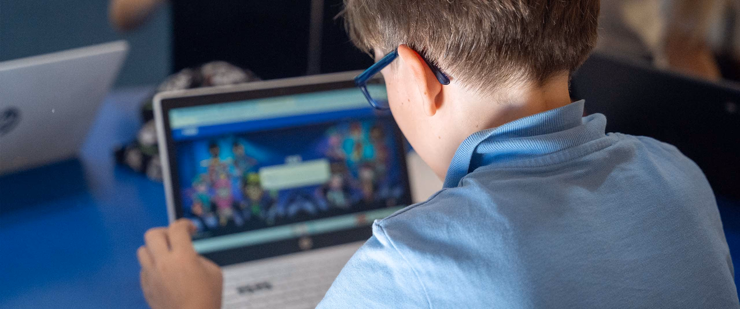 Child wearing blue shirt and glasses using a laptop at a table with others in background.