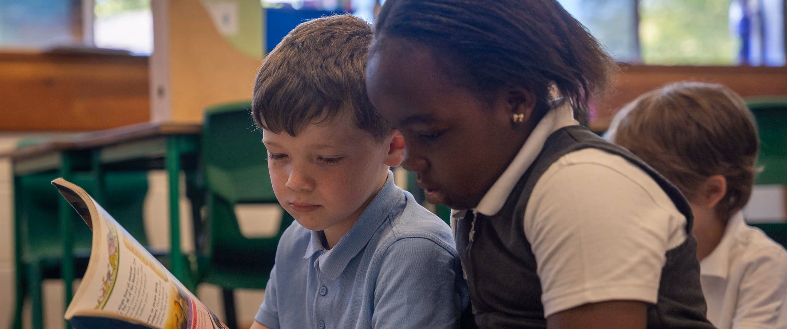 Two young children sitting closely and reading a book together in a classroom.