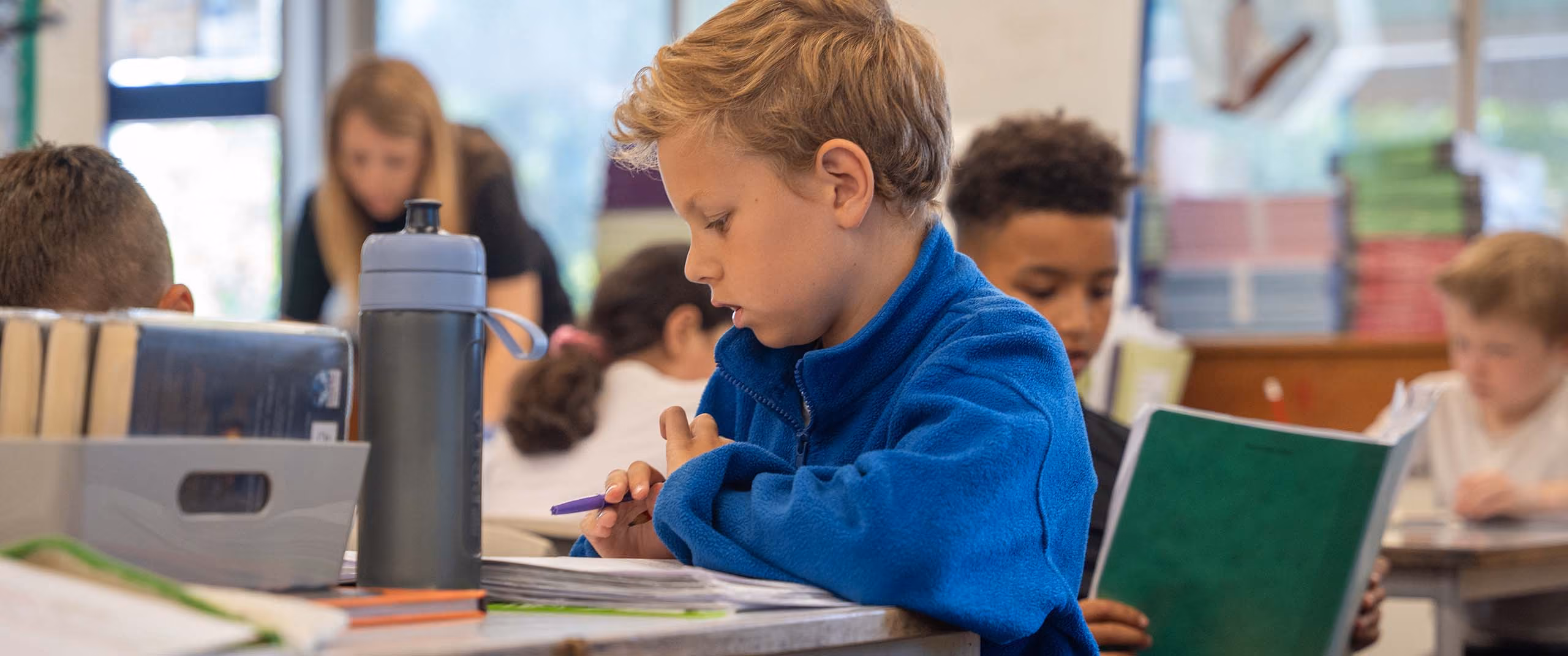 Pupil in a blue fleece holding a pen and reading at his desk, with classmates and a teacher behind.