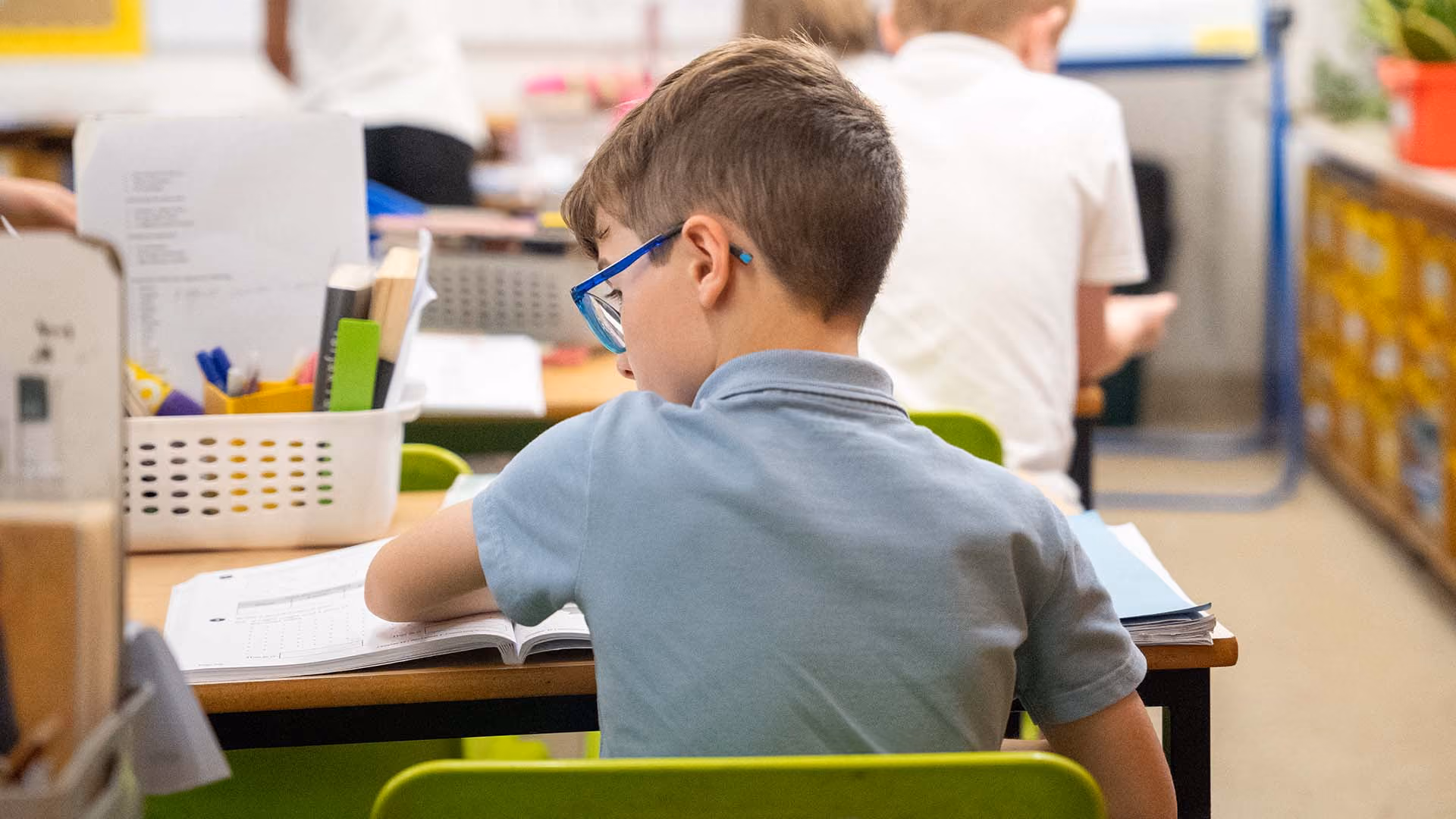Pupil in glasses seen from behind, working from a textbook at his desk.