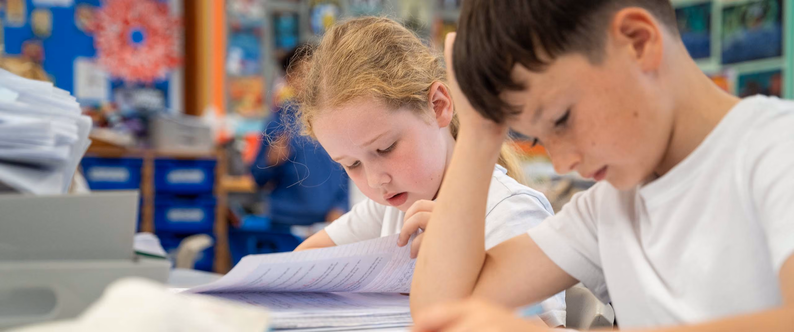 Two pupils concentrating on their exercise books at a classroom desk.