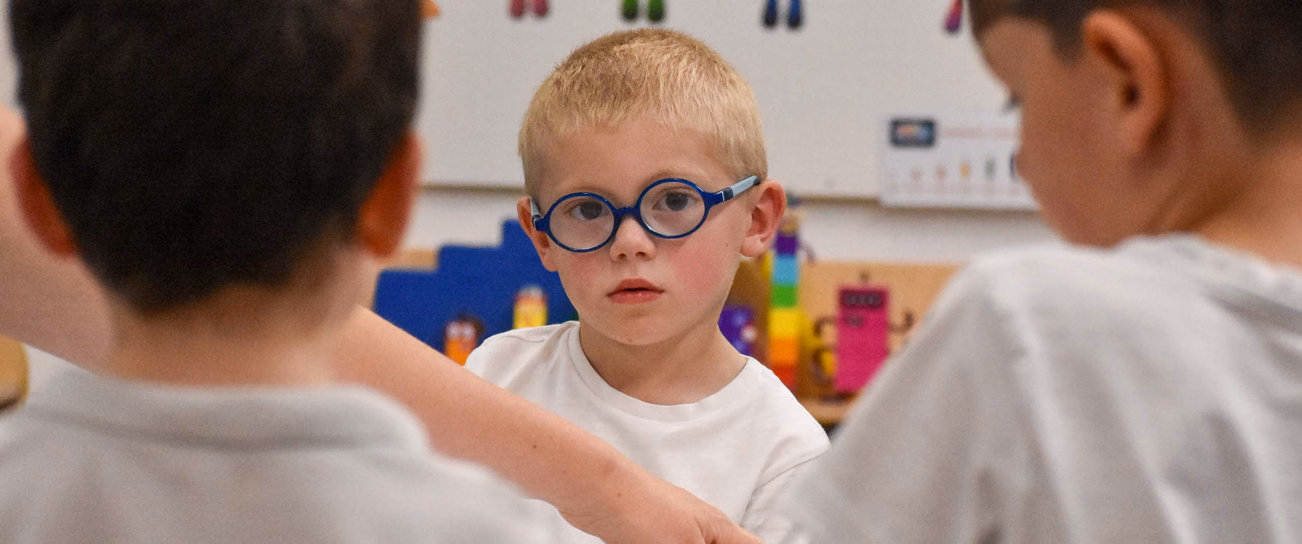 Young pupil in blue glasses looking up from a table, with two pupils seen from behind in the foreground.