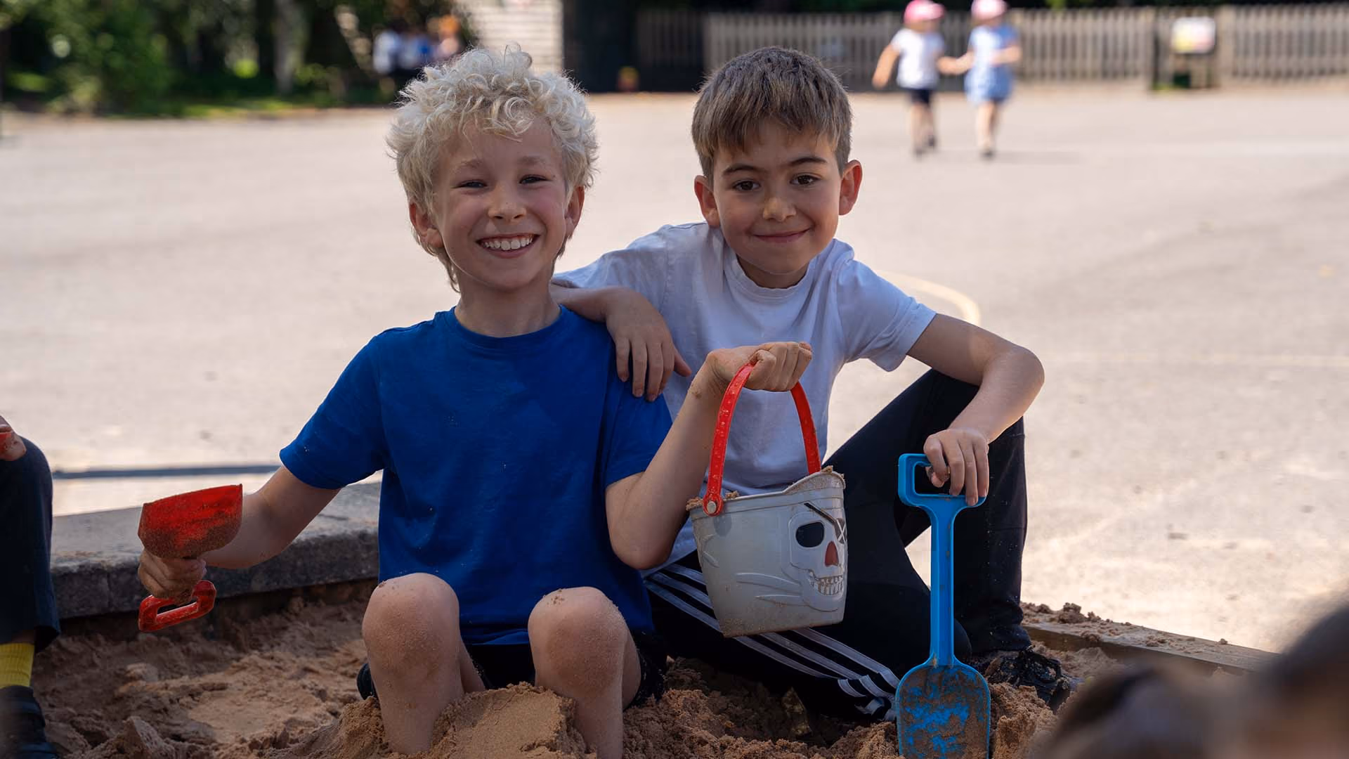 Two boys sitting and smiling in a sandbox holding a skull bucket and blue shovel, outdoors on a sunny day.