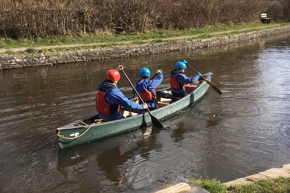 Three pupils in helmets and life jackets paddling a canoe along a canal.