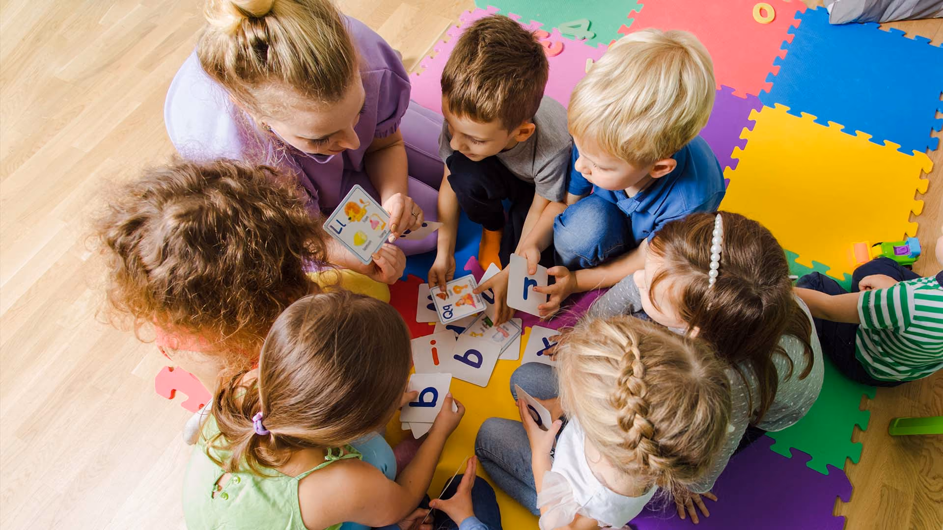 Overhead view of nursery staff and young children playing with letter cards on a colourful floor mat.