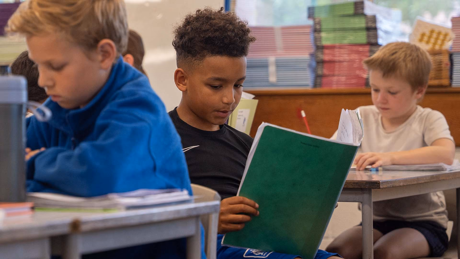 Pupil reading from a green exercise book at his desk, with other pupils working beside him.”