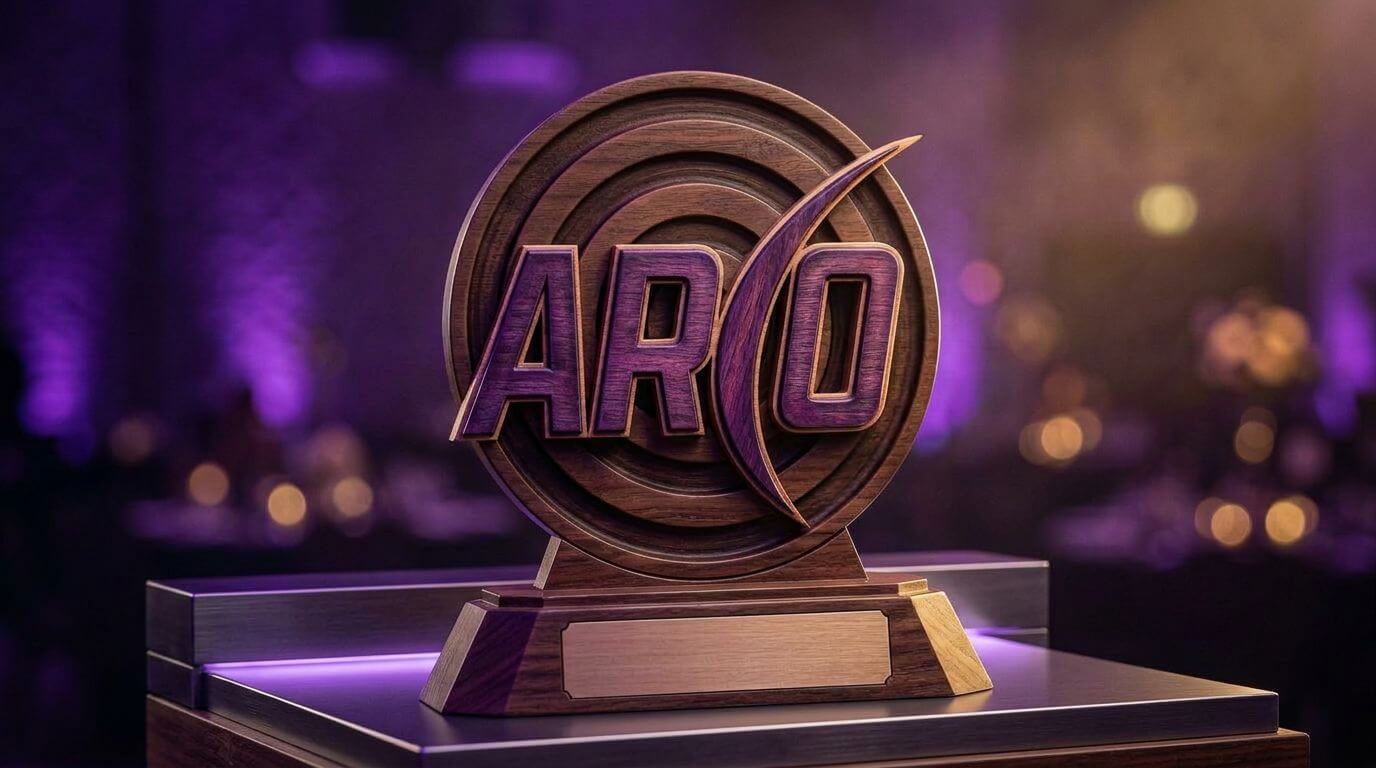 Wooden trophy in front of a blurred purple-lit background, featuring a target design with the word 'ARCO' in bold letters topped by a curved shape resembling an arrow.