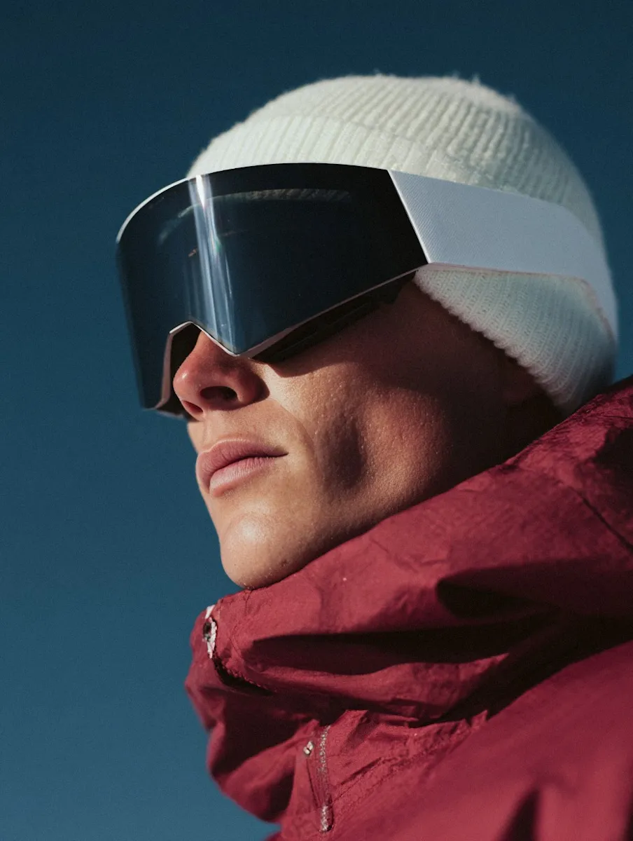 Close-up of a person wearing a white knit beanie, large black ski goggles, and a red winter jacket against a clear blue sky.