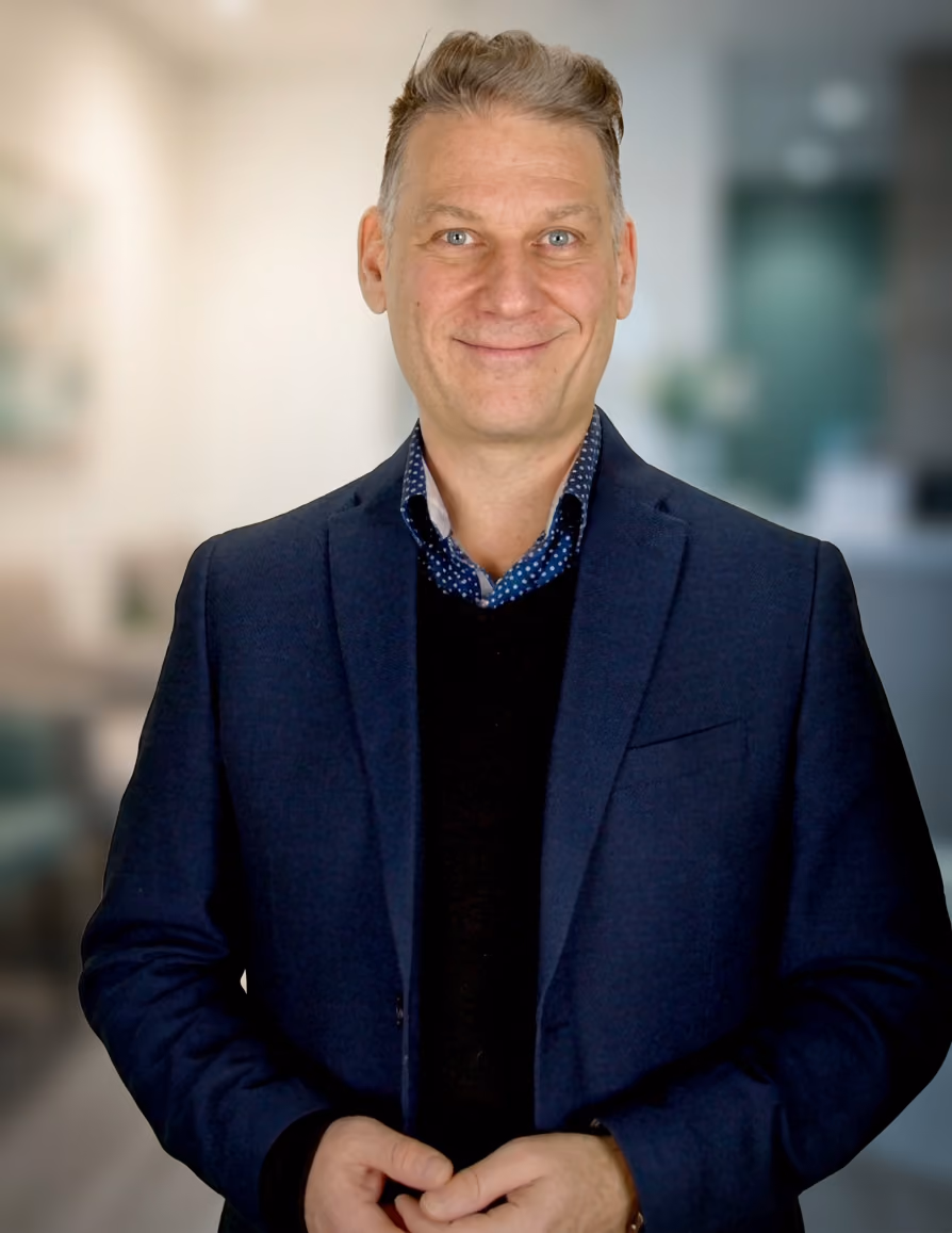 Portrait d'un homme souriant aux cheveux gris portant un veston bleu marine et une chemise à motif pointillé bleu, en arrière-plan flou d'un intérieur moderne.