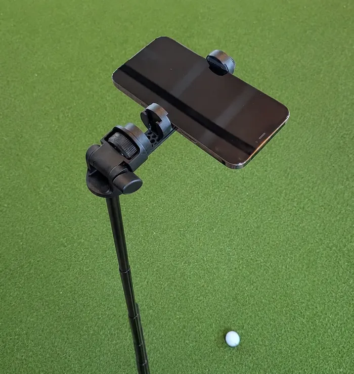 A smartphone mounted on a small tripod stand aimed at a golf ball resting on artificial turf.