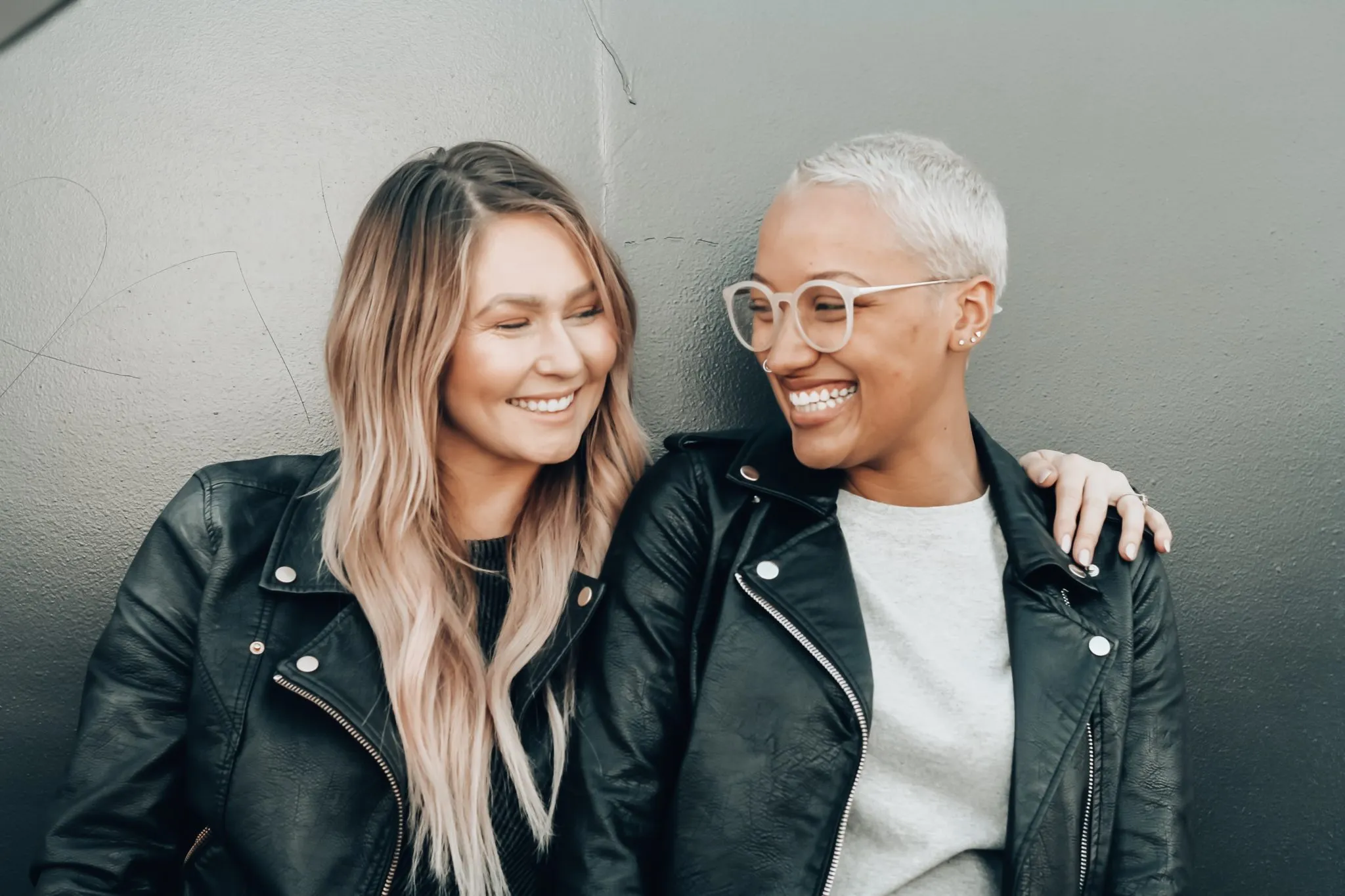 Two smiling women in leather jackets laughing against a gray wall.