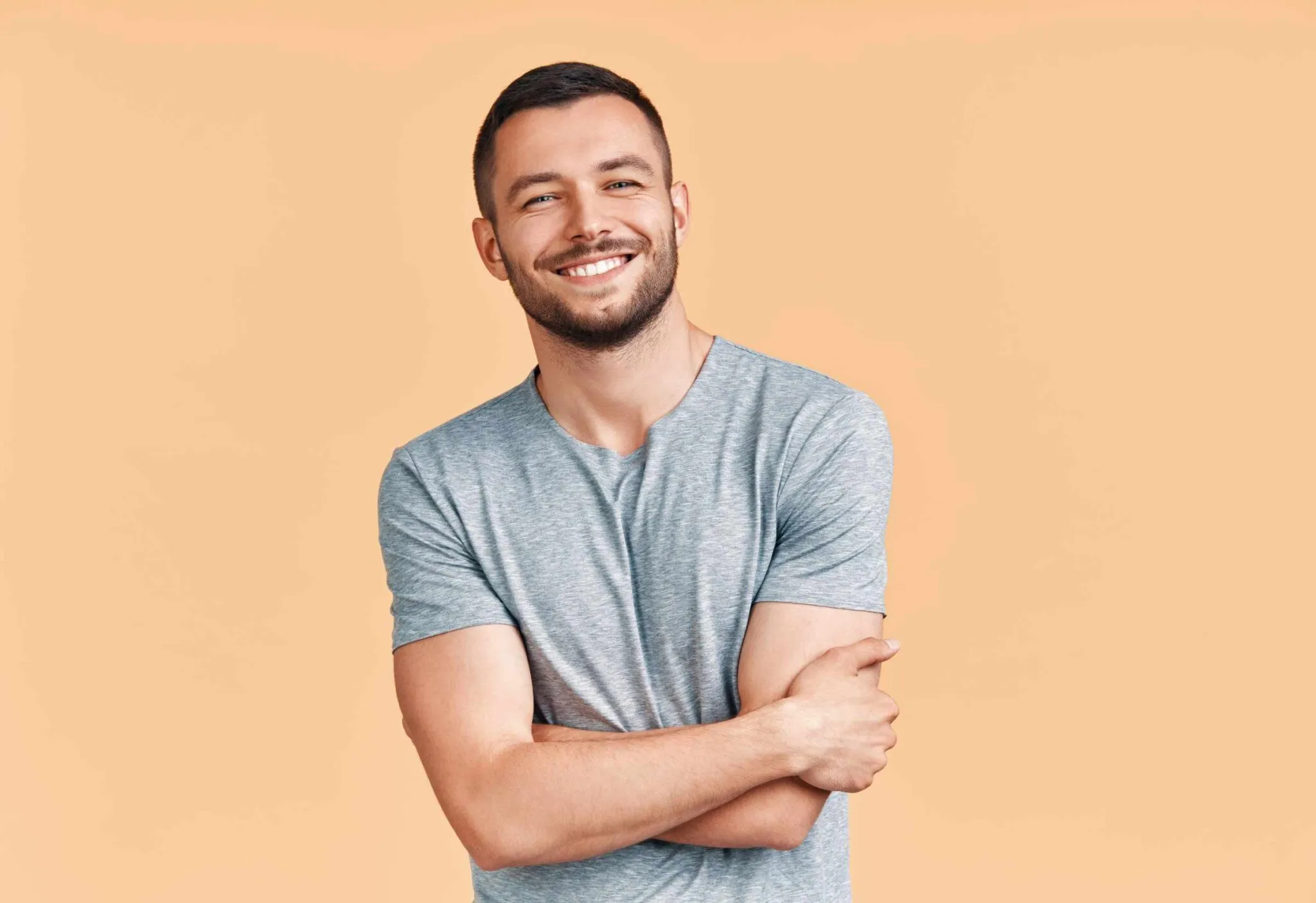 Smiling man in a gray T-shirt with arms crossed against a peach background.